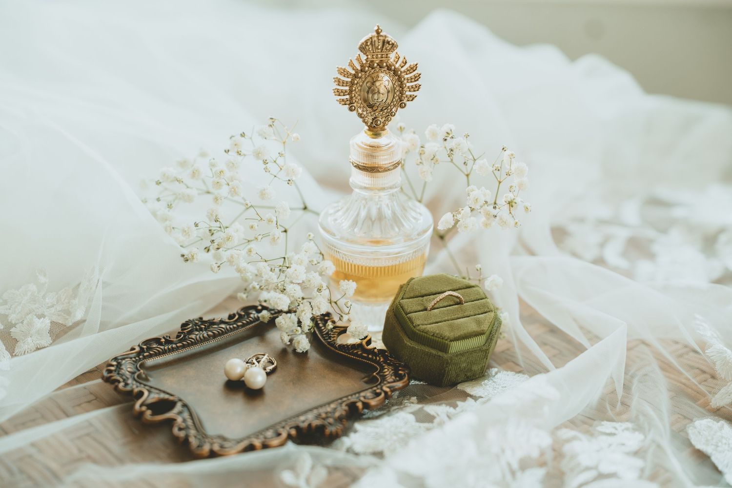 Vintage wedding flat lay featuring ornate perfume bottle, green velvet ring box, pearl earrings, and baby breath flowers.