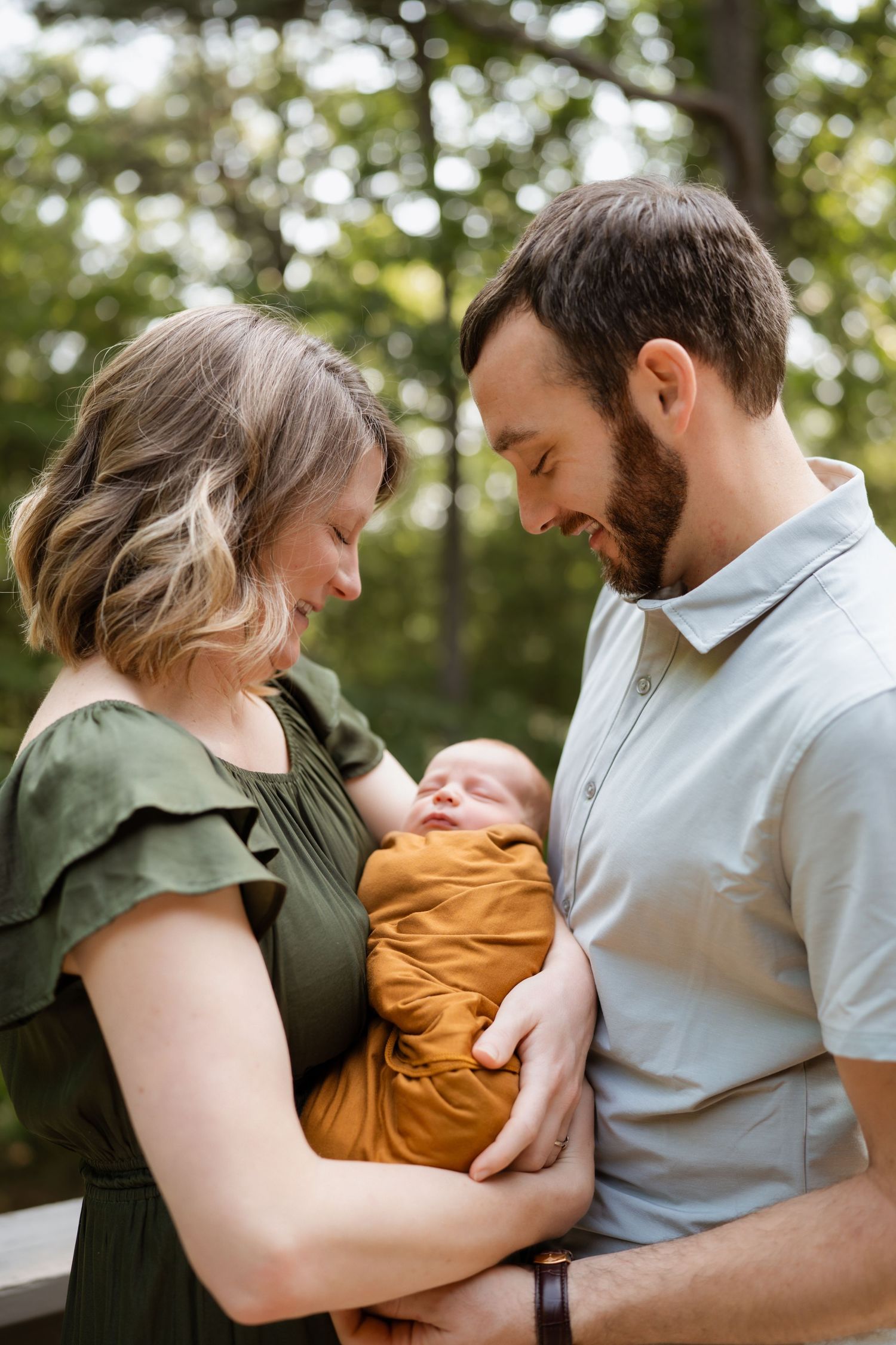 New parents gazing lovingly at their newborn wrapped in a mustard swaddle outdoors.