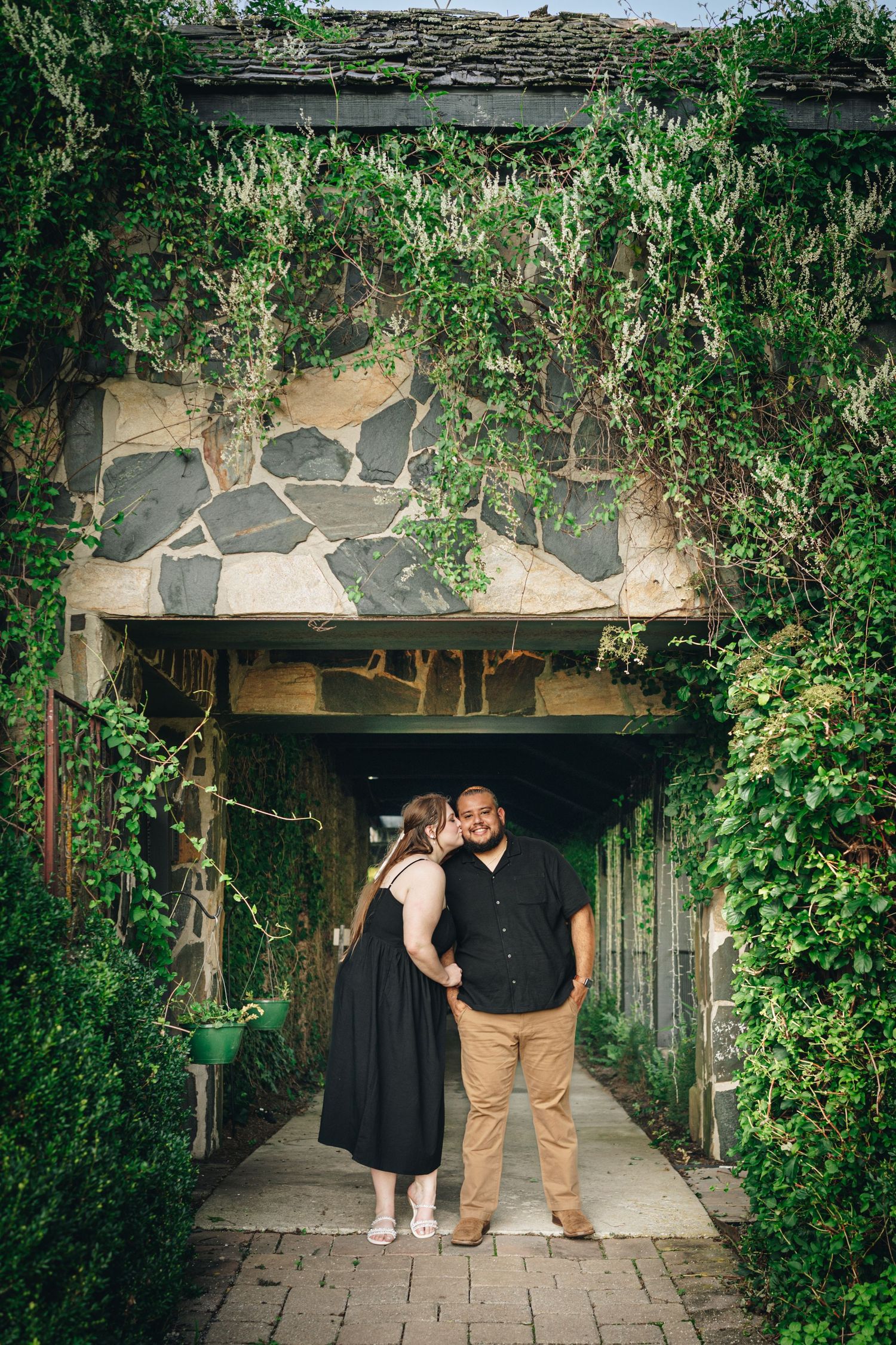 Couple sharing a kiss under an ivy-covered stone archway entrance surrounded by lush green foliage.