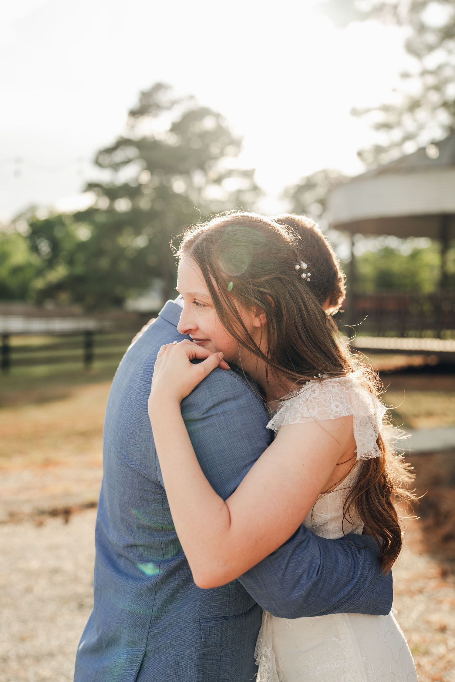Bride in lace wedding dress embracing groom in blue suit during golden hour outdoor wedding photos.