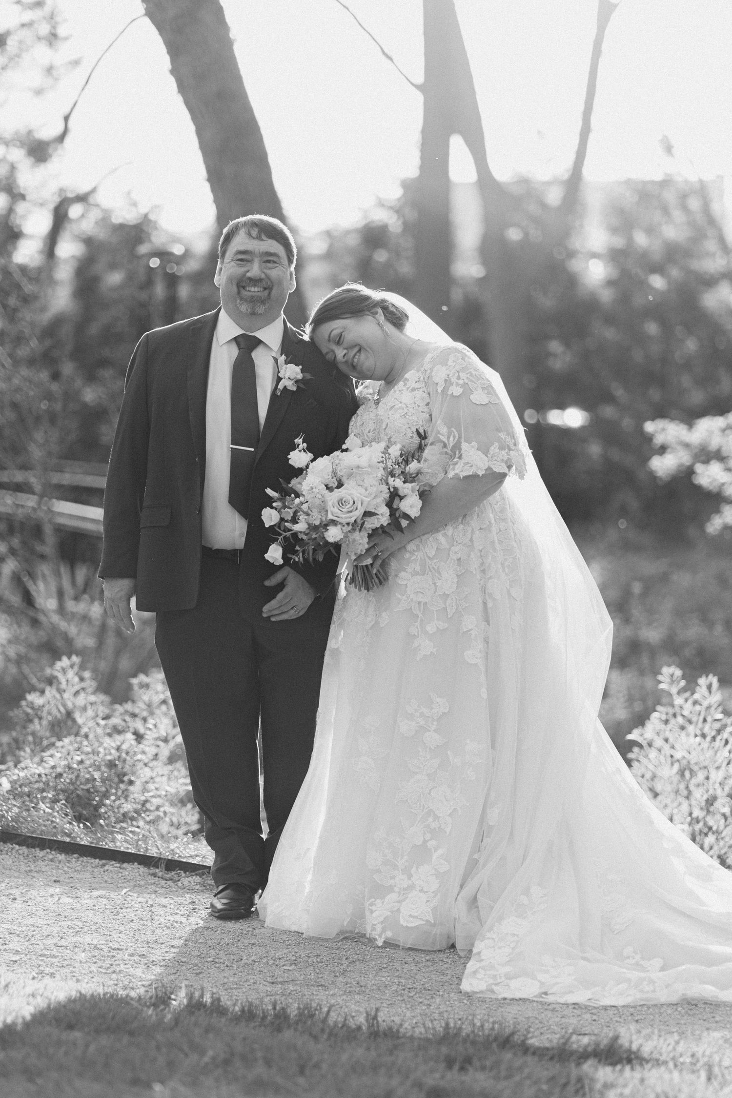 Black and white portrait of a smiling bride and groom posing together outdoors holding a floral bouquet.