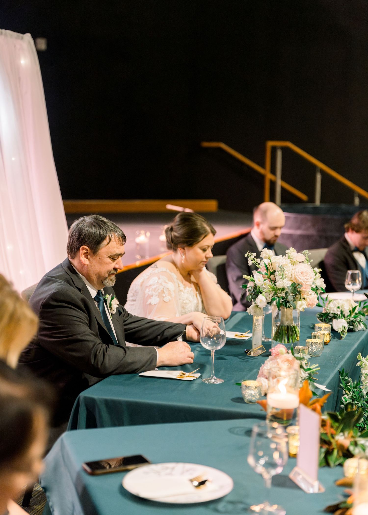 Bride and groom seated at teal-draped sweetheart table with floral centerpieces at wedding reception.