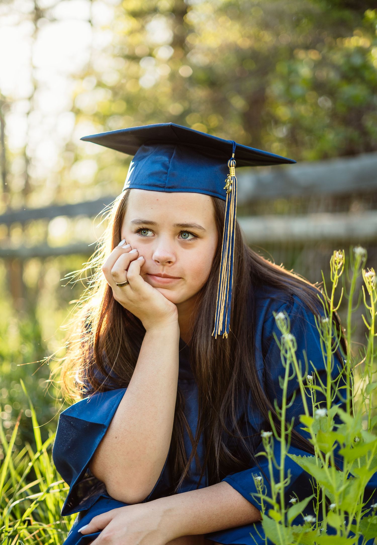 A brunette girl in a blue cap and gown rests her chin on her hand while sitting in a sunlit meadow.