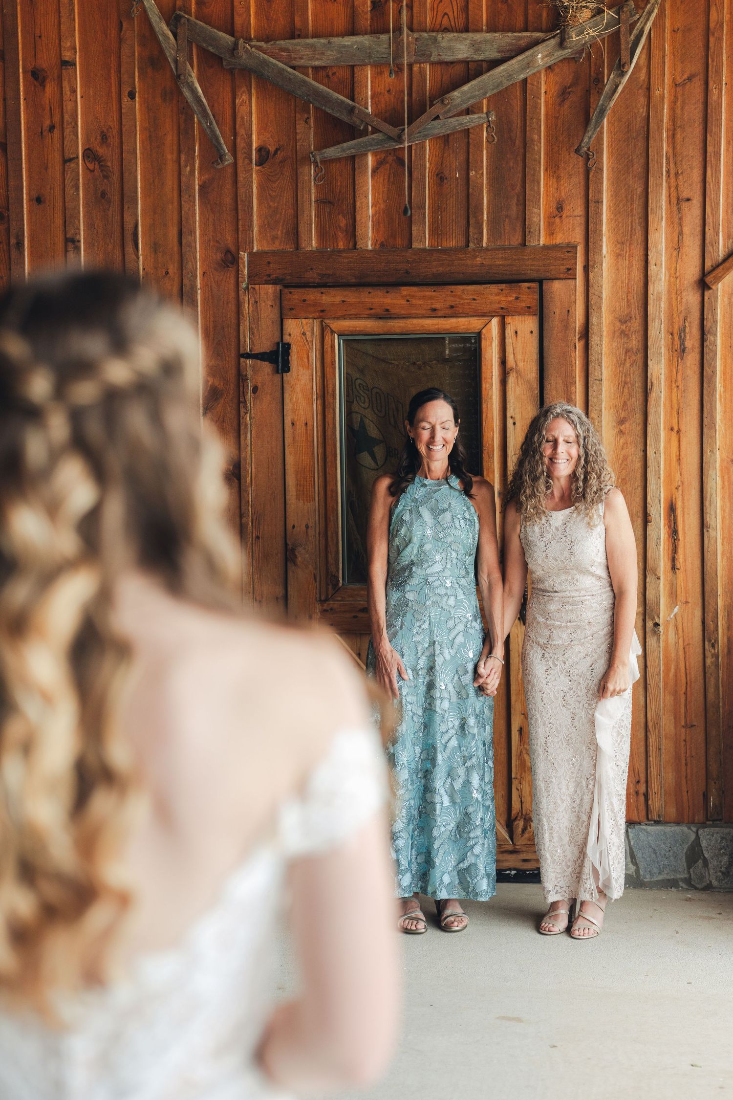 Two women in elegant gowns smile at a bride during a rustic barn wedding getting-ready moment.