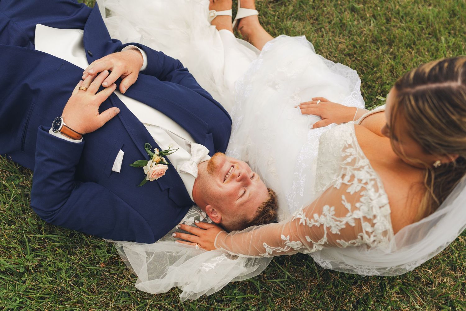 Bride and groom laughing joyfully while lying on green grass in wedding attire, captured from above.