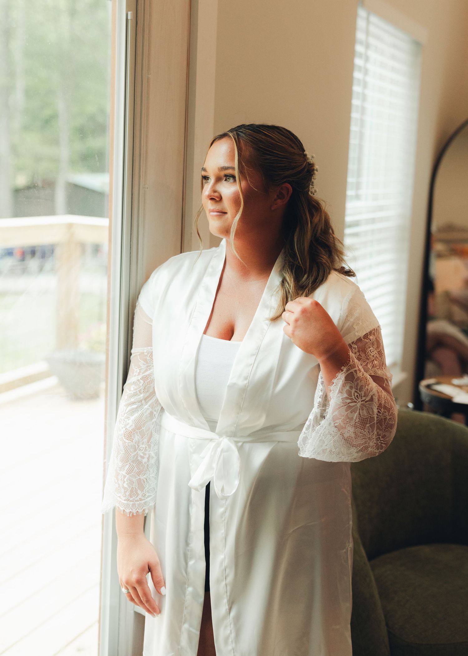 Bride in white lace robe gazing out window during wedding morning preparation.