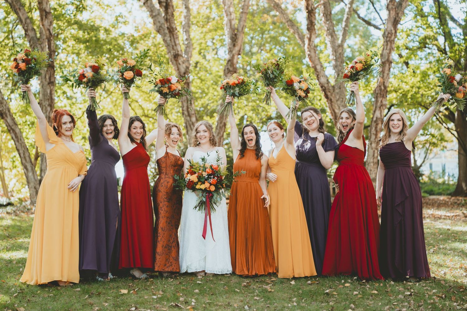 Bridesmaids in mismatched fall-colored gowns holding bouquets under trees in autumn.