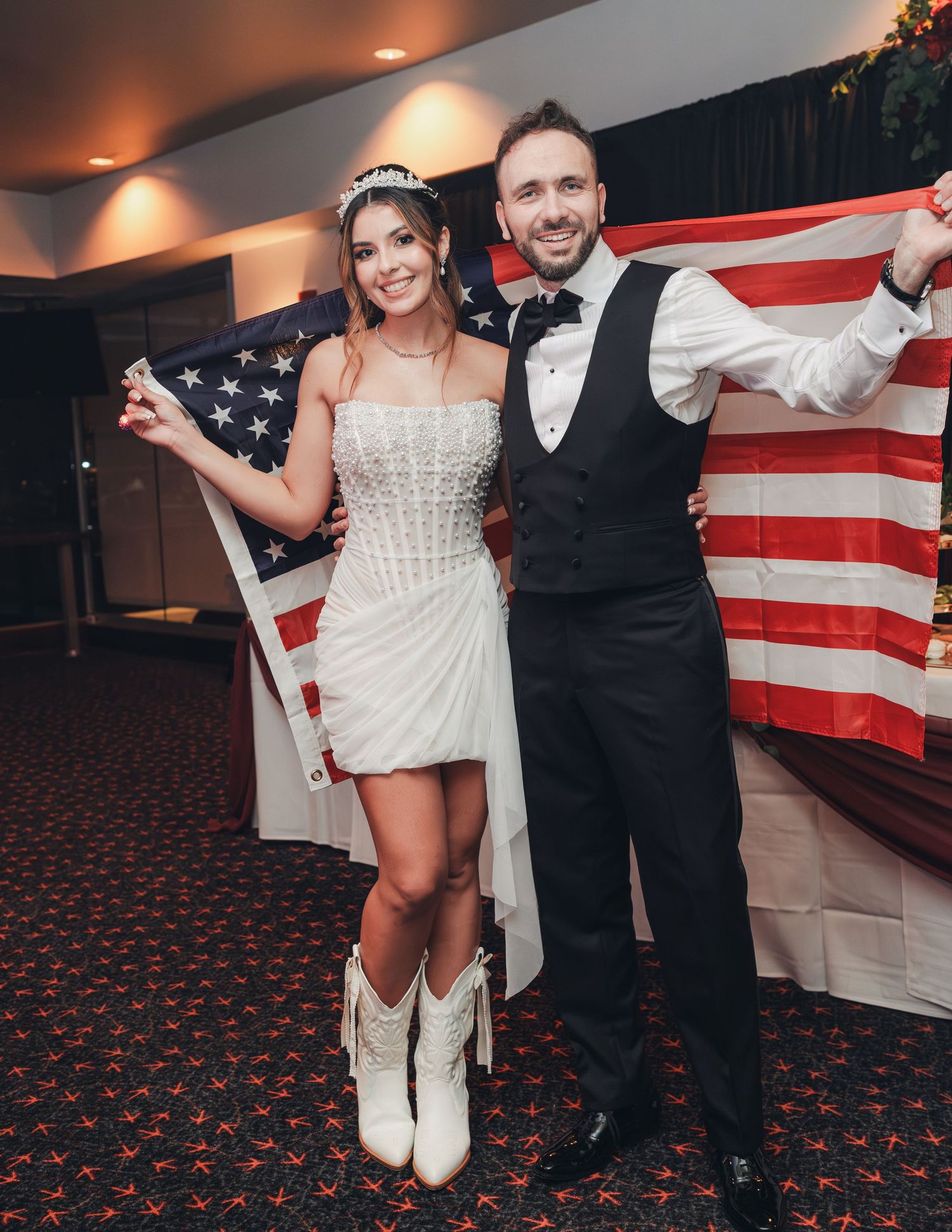 Bride in white mini dress and cowboy boots poses with groom holding American flag at wedding reception.