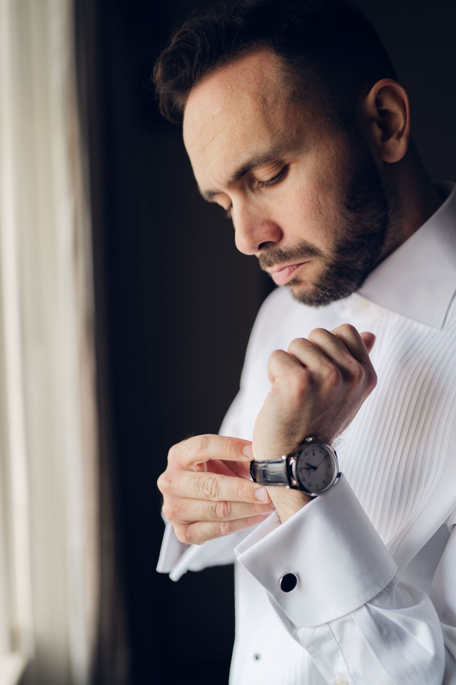 Handsome groom in a white dress shirt fastening his cufflinks while wearing a classic watch.
