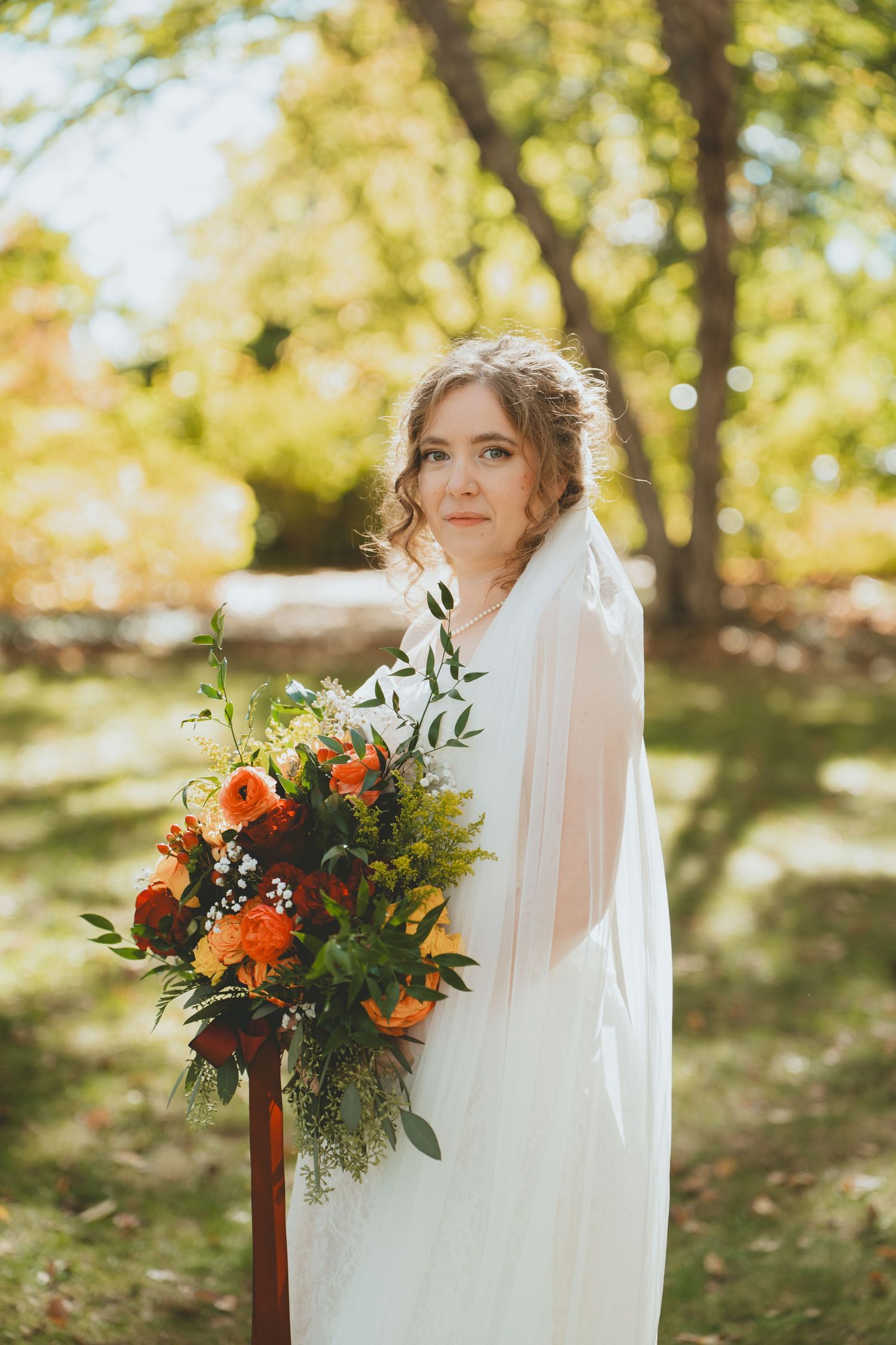 Bride in a white gown and veil holding a large autumn bouquet of orange and red flowers outdoors.