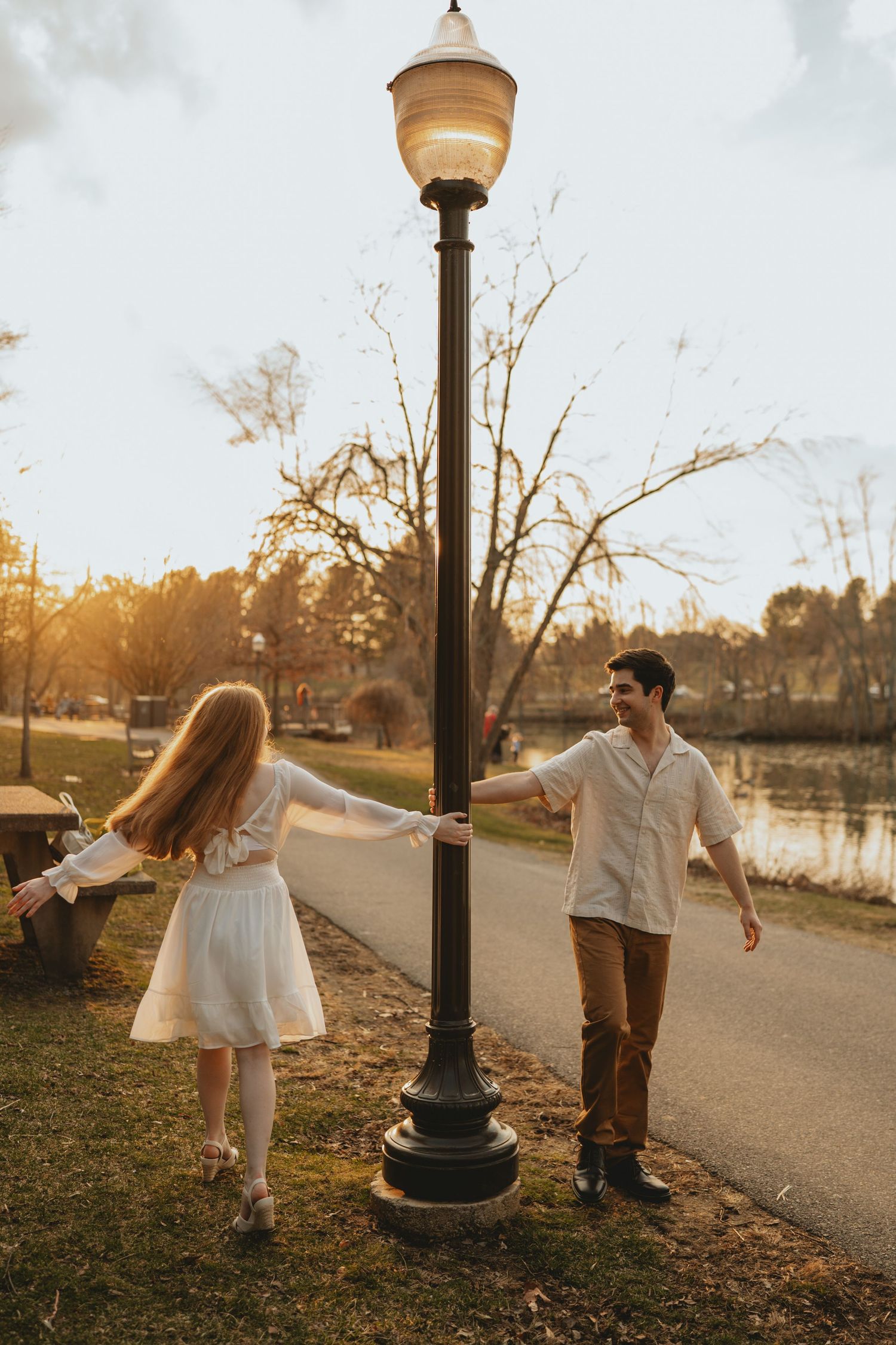 Couple playfully dancing around a glowing lamppost in a sunlit park during golden hour engagement session.
