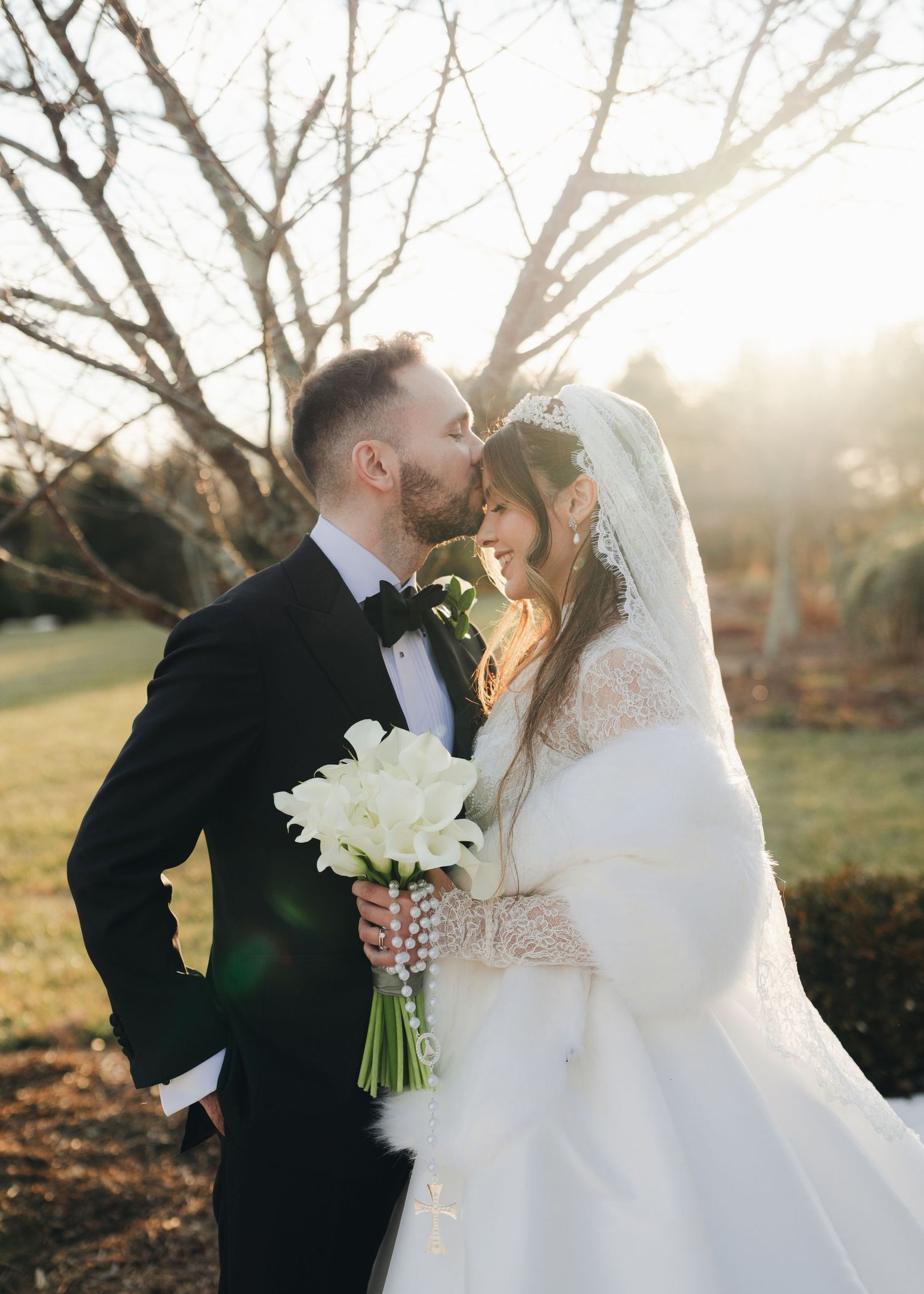 Bride and groom share a romantic kiss outdoors in golden sunset light, bride holding white calla lily bouquet.