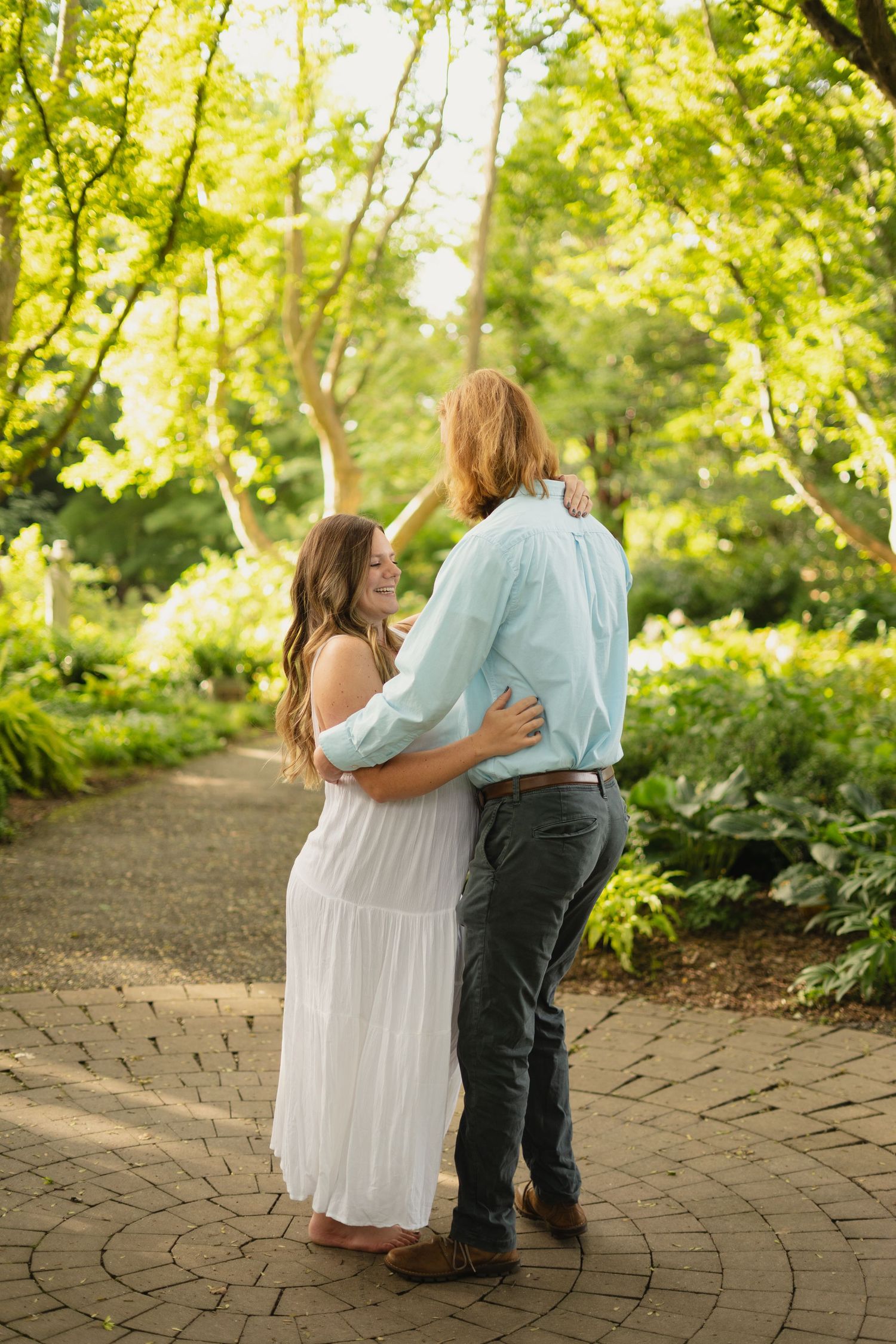 Pregnant woman in white dress embracing partner on sunlit garden brick path.