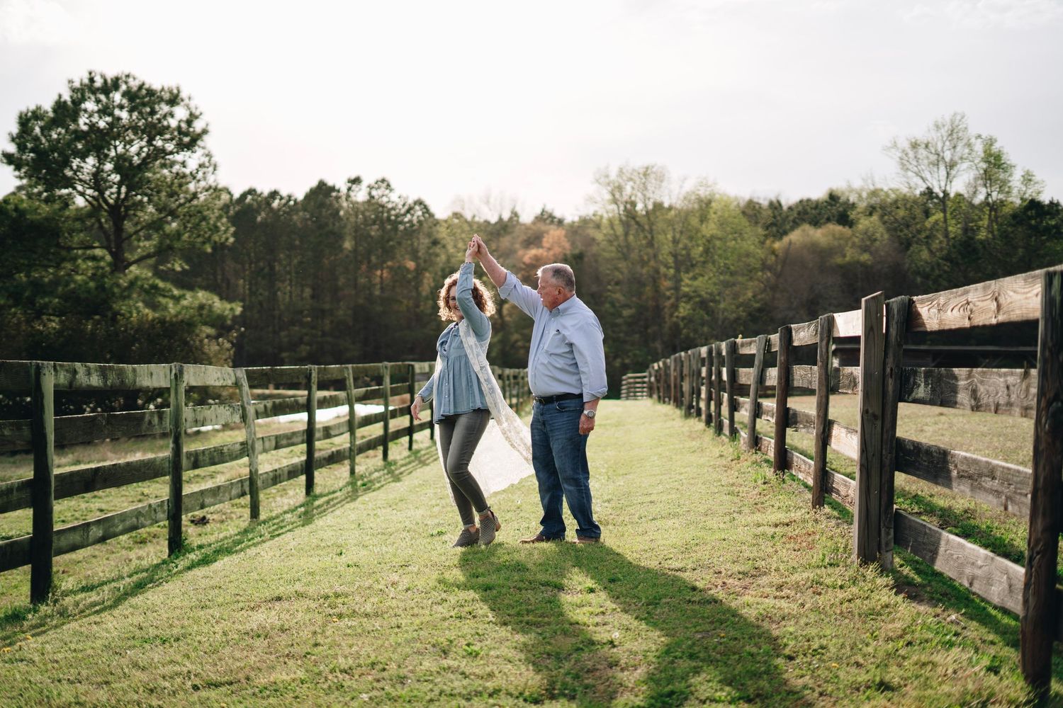 Couple dancing joyfully between wooden farm fences on green grass with trees in background.