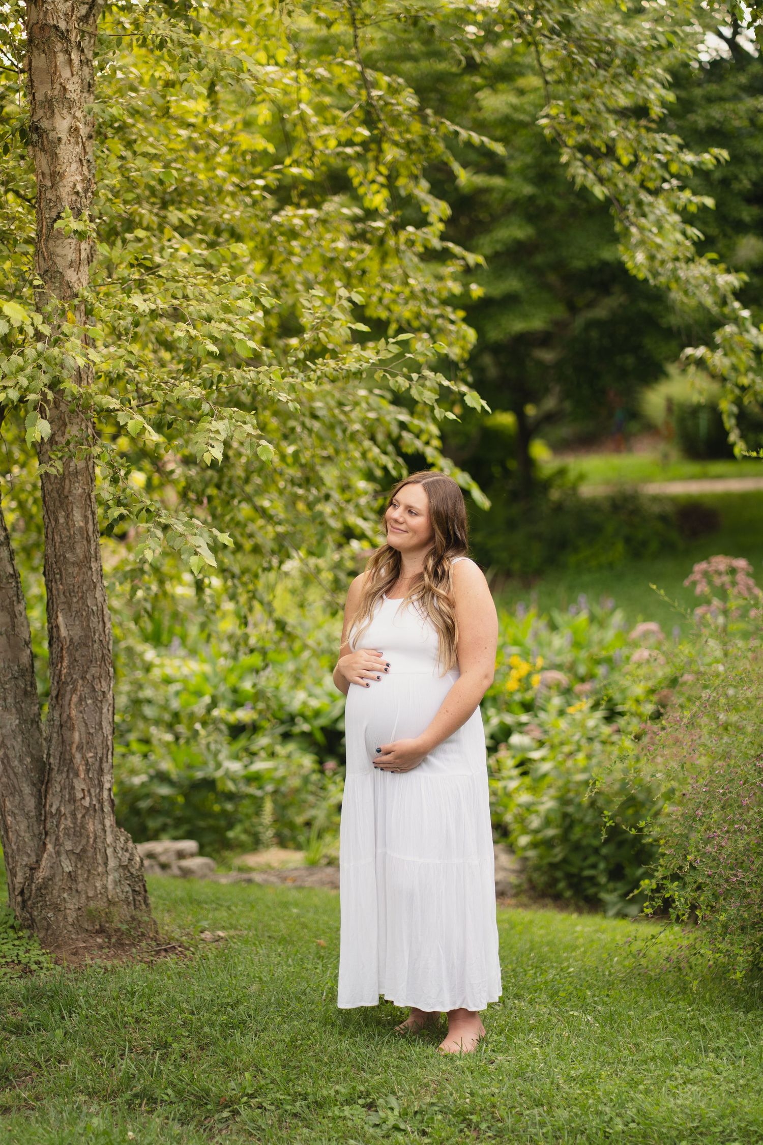 Pregnant woman in a white maxi dress cradling her baby bump in a lush green garden setting.