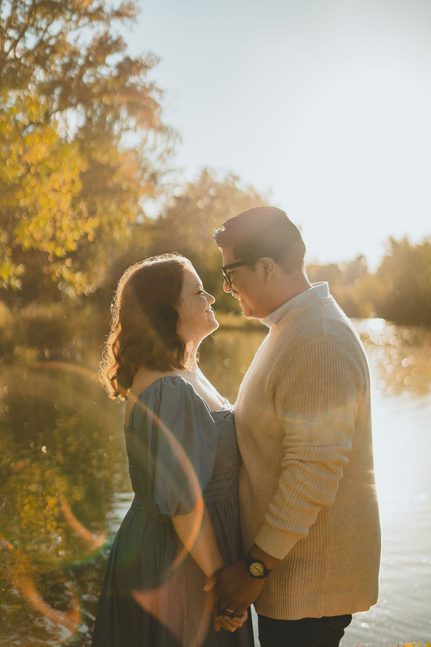 Couple holding hands and gazing at each other by a sunlit lake.