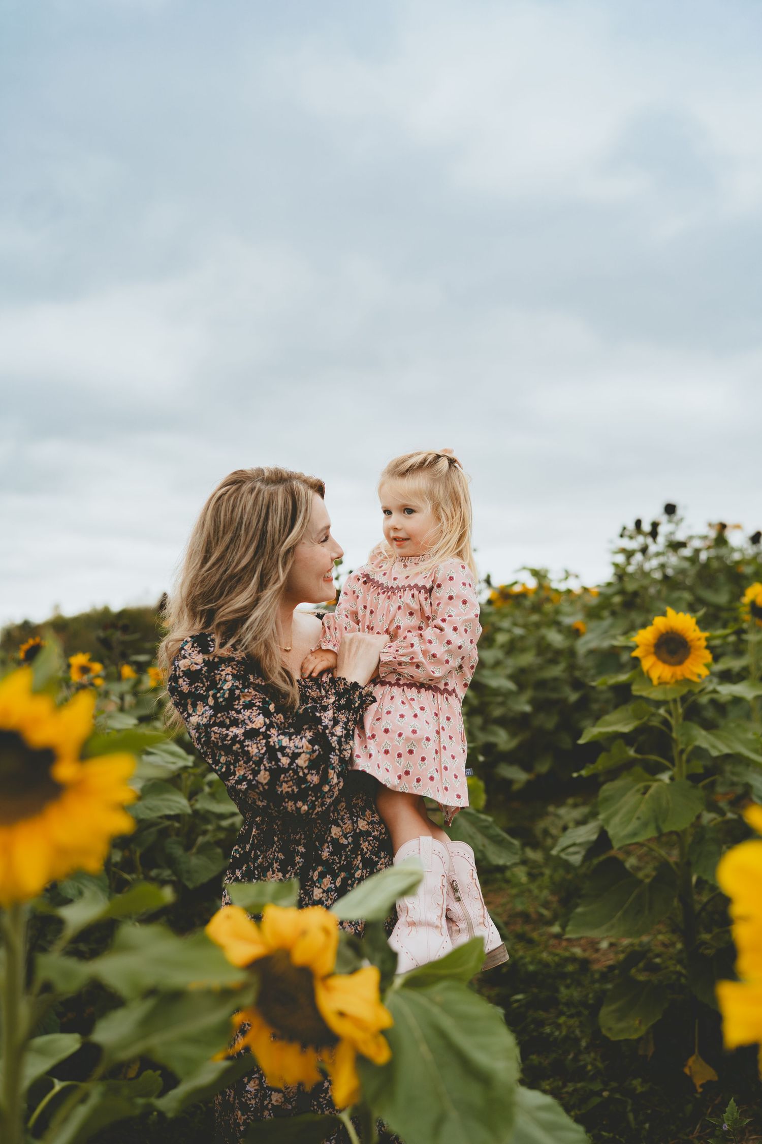 Blonde mother lifting little girl in pink dress among sunflowers.