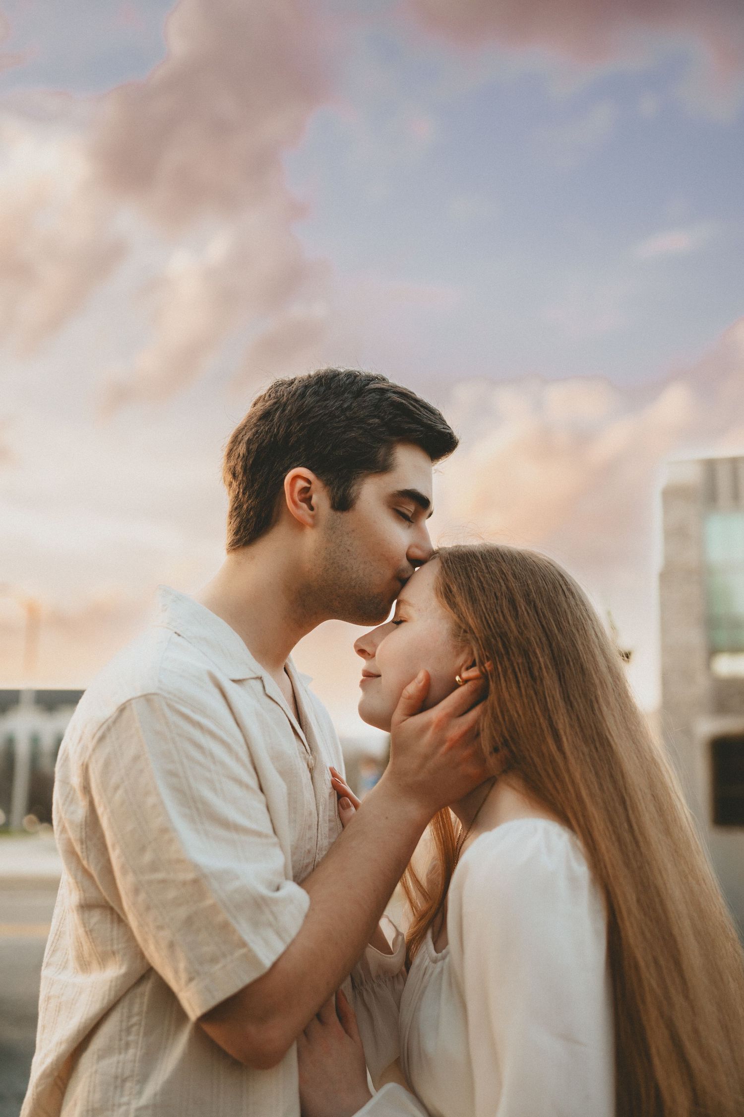 Couple sharing a tender kiss against a pastel sunset sky, man gently cupping womans face.