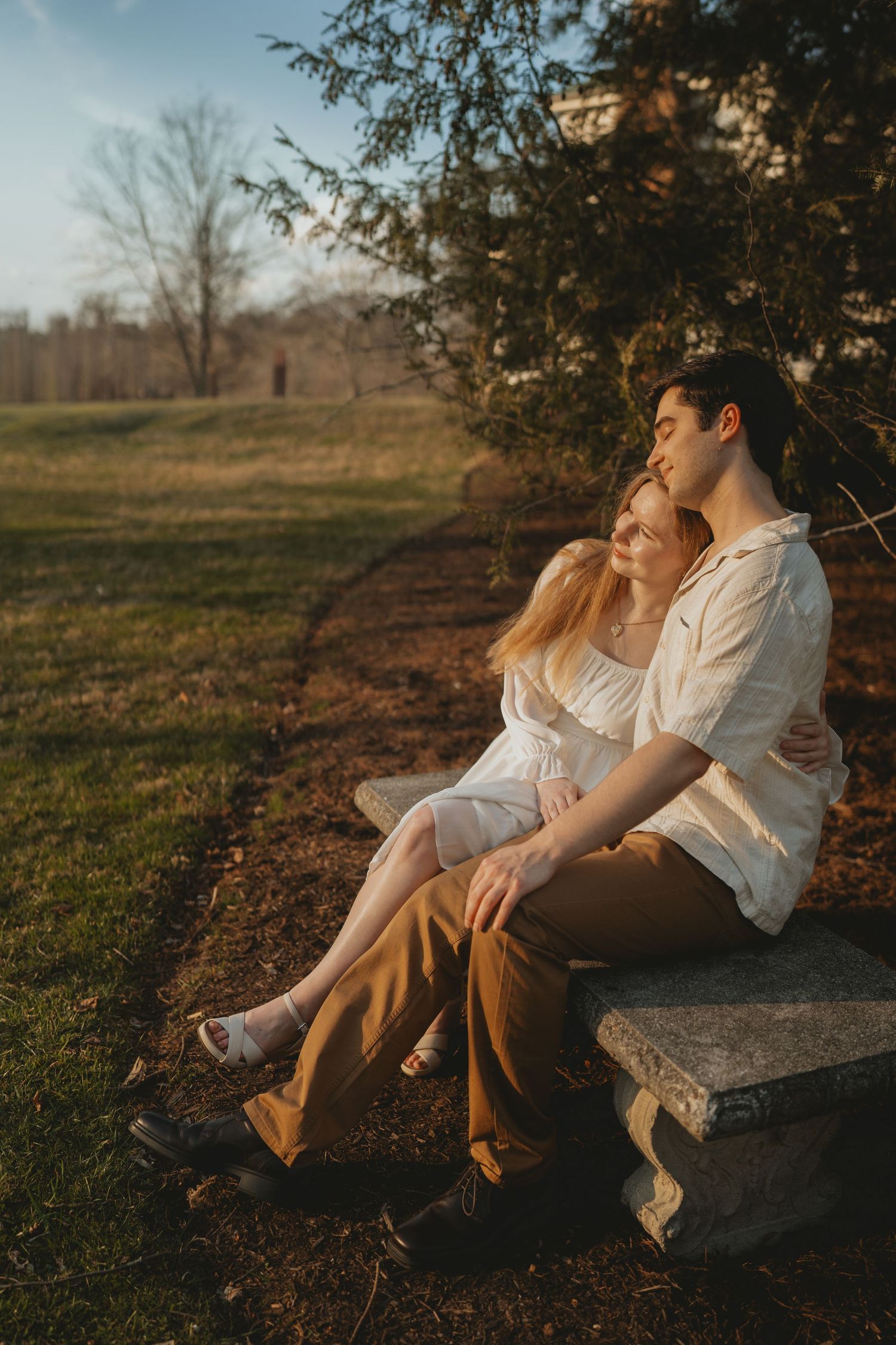 A couple sits closely on a stone bench at golden hour, embracing peacefully in a scenic park setting.