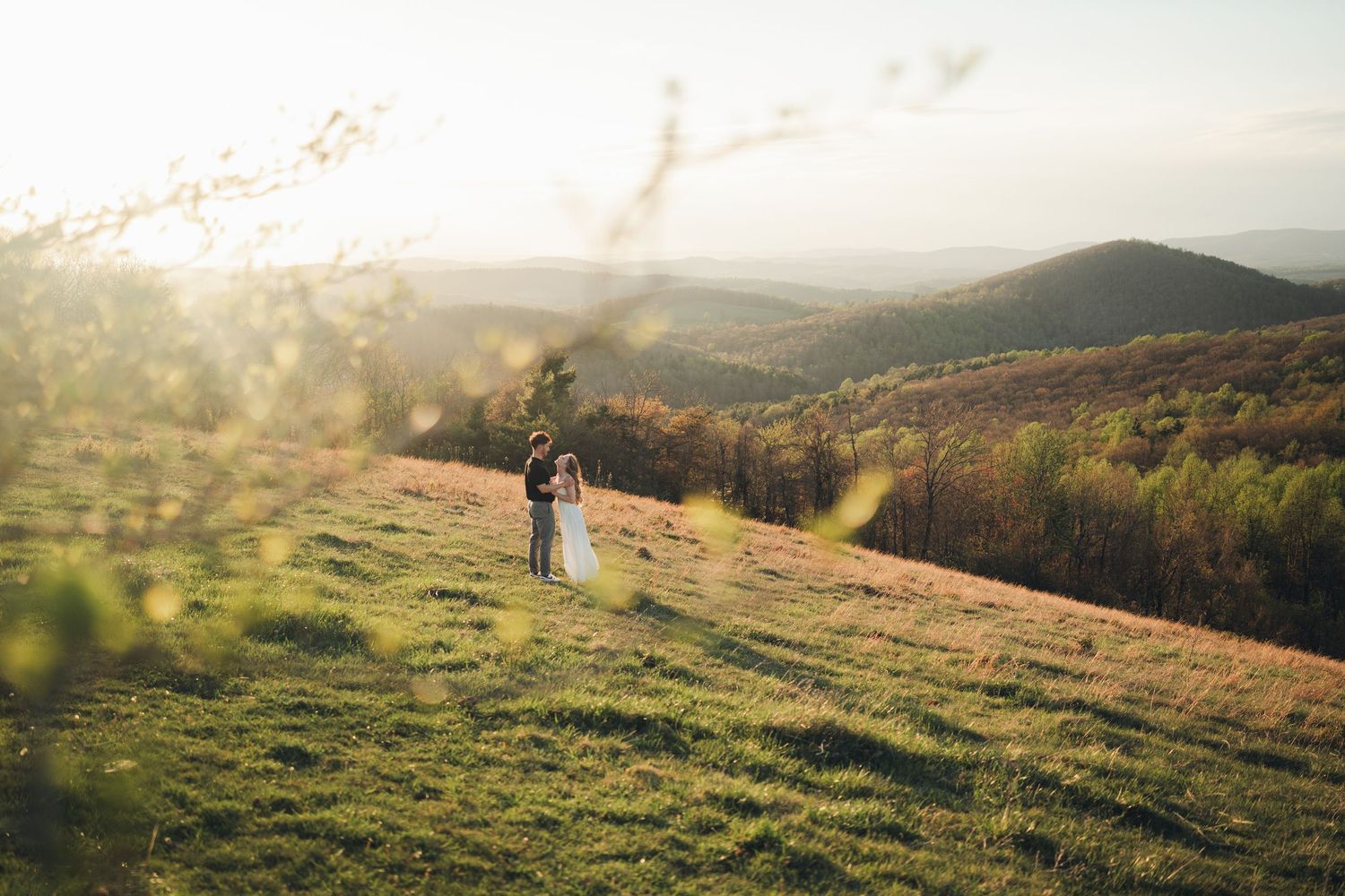 Romantic wedding couple embraces on golden hilltop meadow with sweeping autumn mountain landscape behind them.