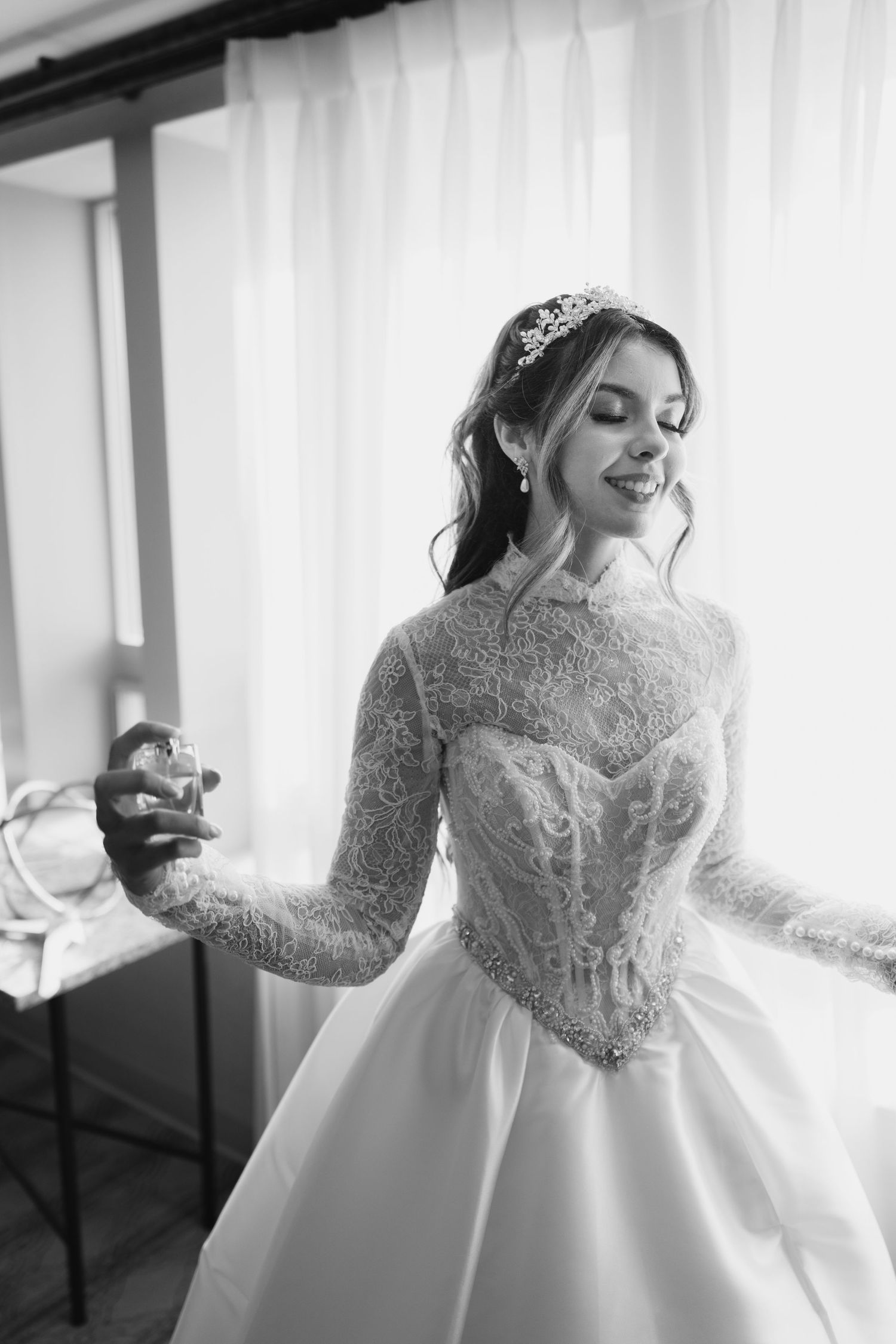 Black and white portrait of smiling bride in lace gown and tiara holding perfume bottle by window.