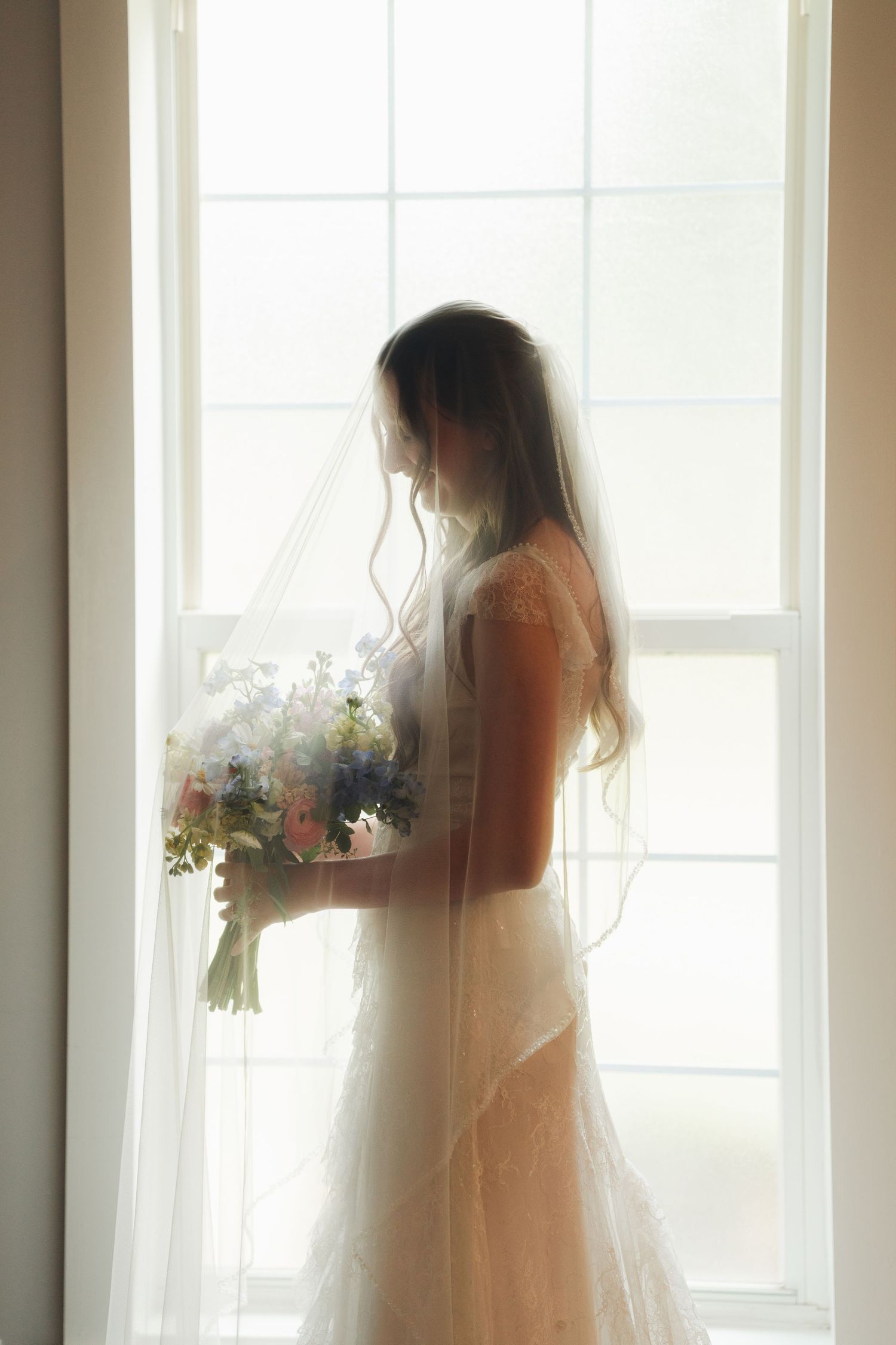 Bride in lace wedding gown and veil holding floral bouquet silhouetted against bright window light.