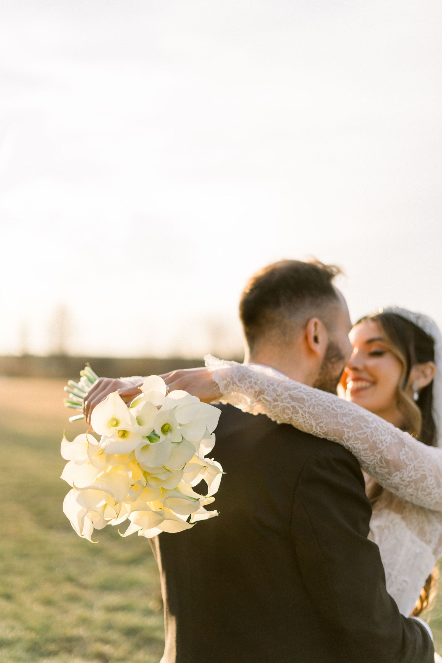 Smiling bride embraces groom holding white calla lily bouquet at golden hour.
