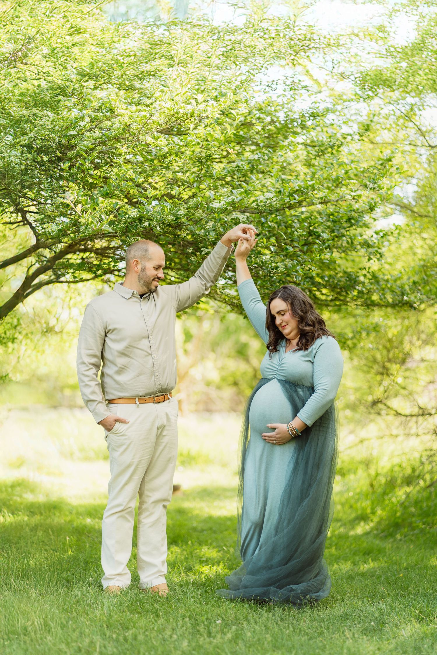 Man spinning pregnant woman in teal tulle gown during outdoor maternity photoshoot under green trees.