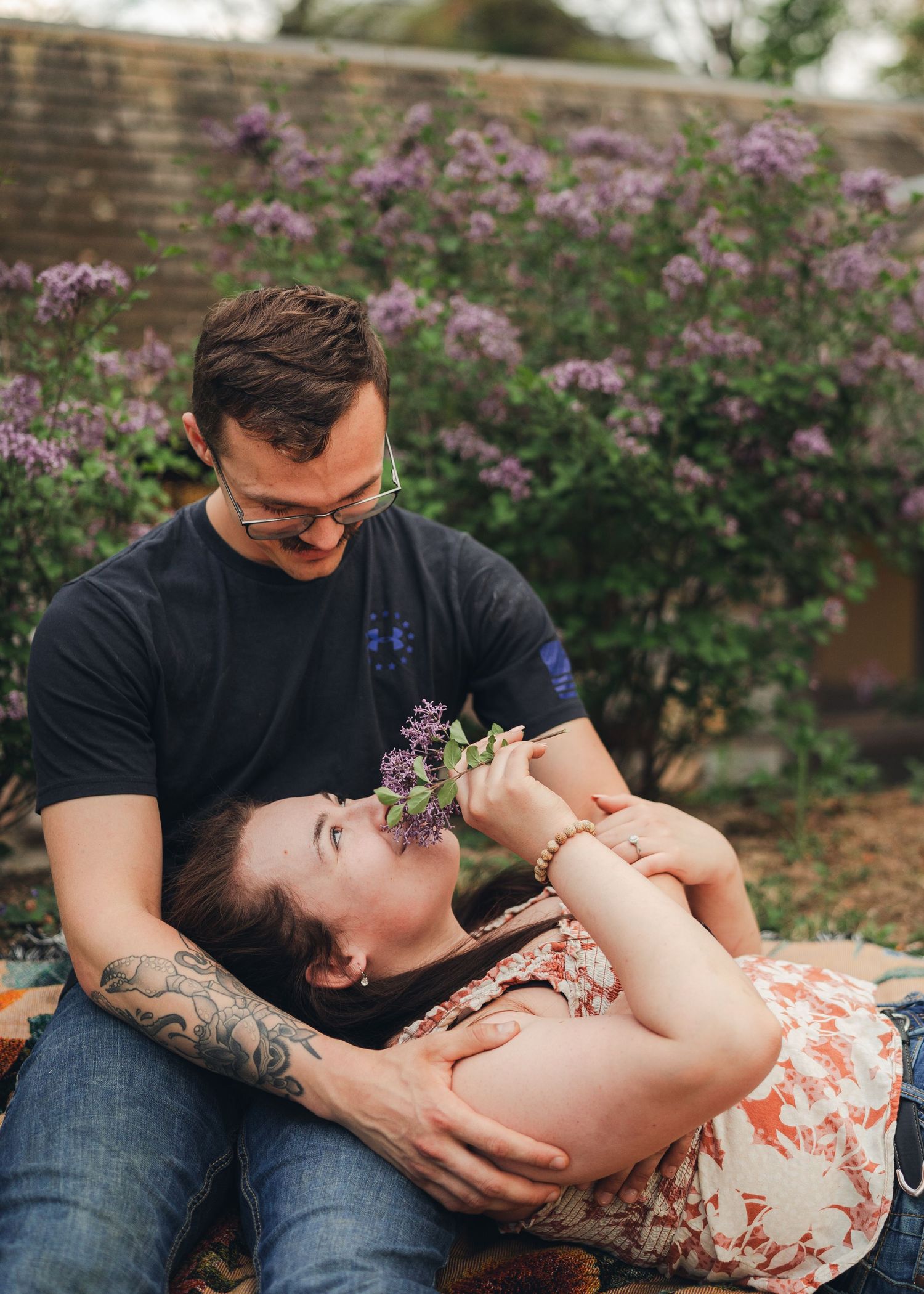 Couple relaxing outdoors with man holding lilac flowers to womans nose by purple bushes.