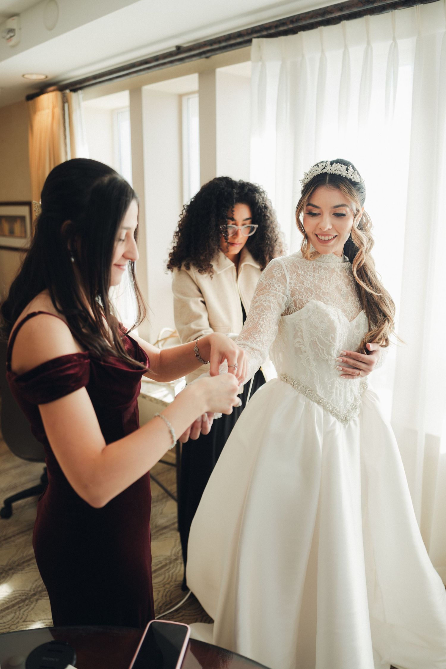 Bride in lace gown and tiara being helped by two bridesmaids while getting ready before wedding.