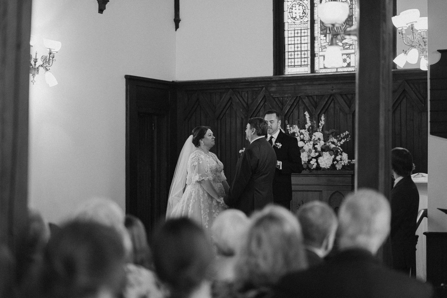 Black and white photo of bride and groom exchanging vows at church altar before seated wedding guests.