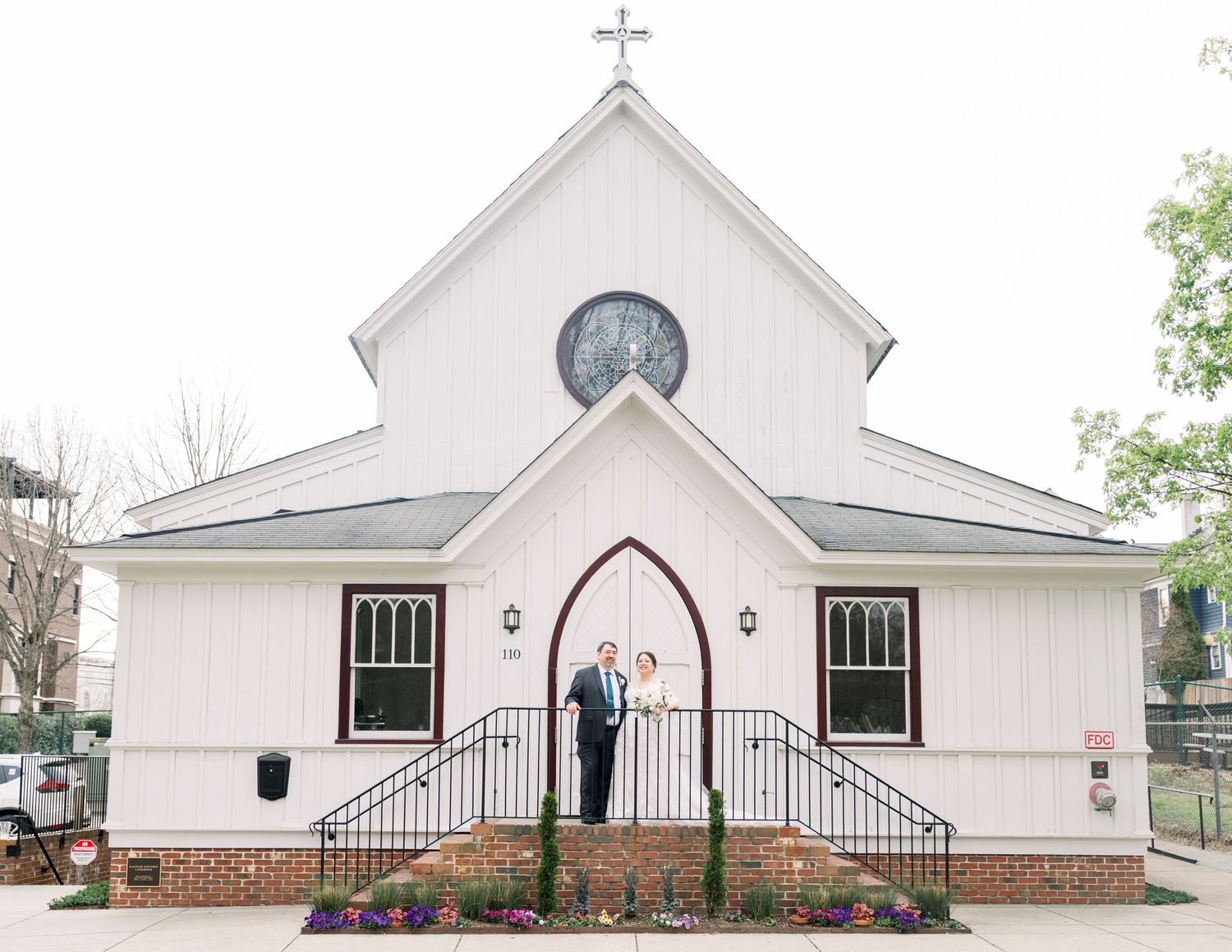 Bride and groom posing on steps of a charming white chapel with a cross and arched doorway.