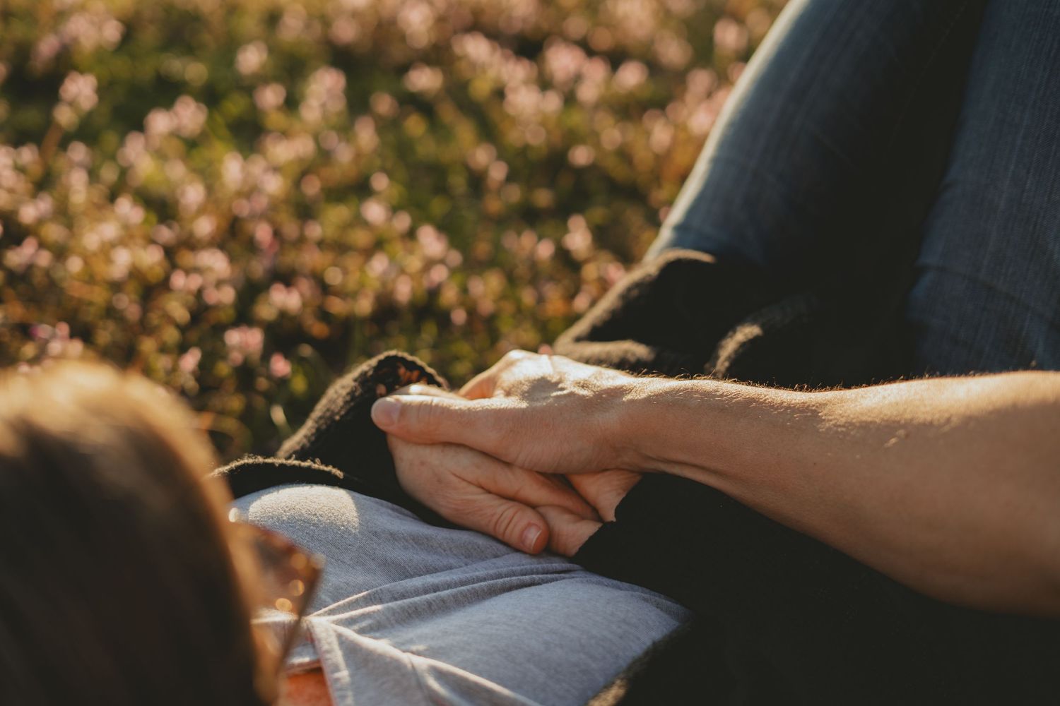 Two people holding hands outdoors in a sunlit field of pink flowers.