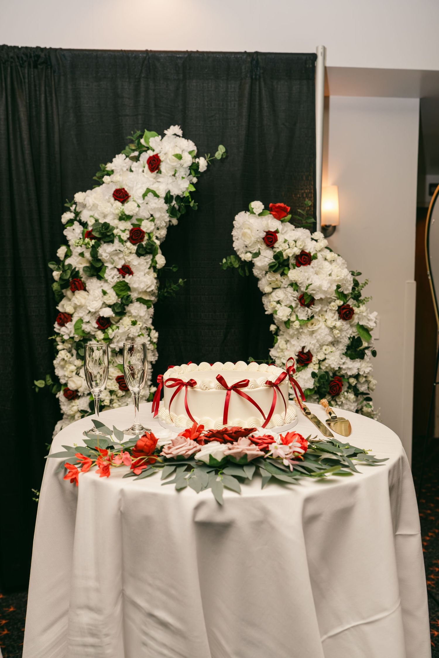 Elegant white wedding cake with red ribbon bows displayed on table with floral arch backdrop of white and red roses.