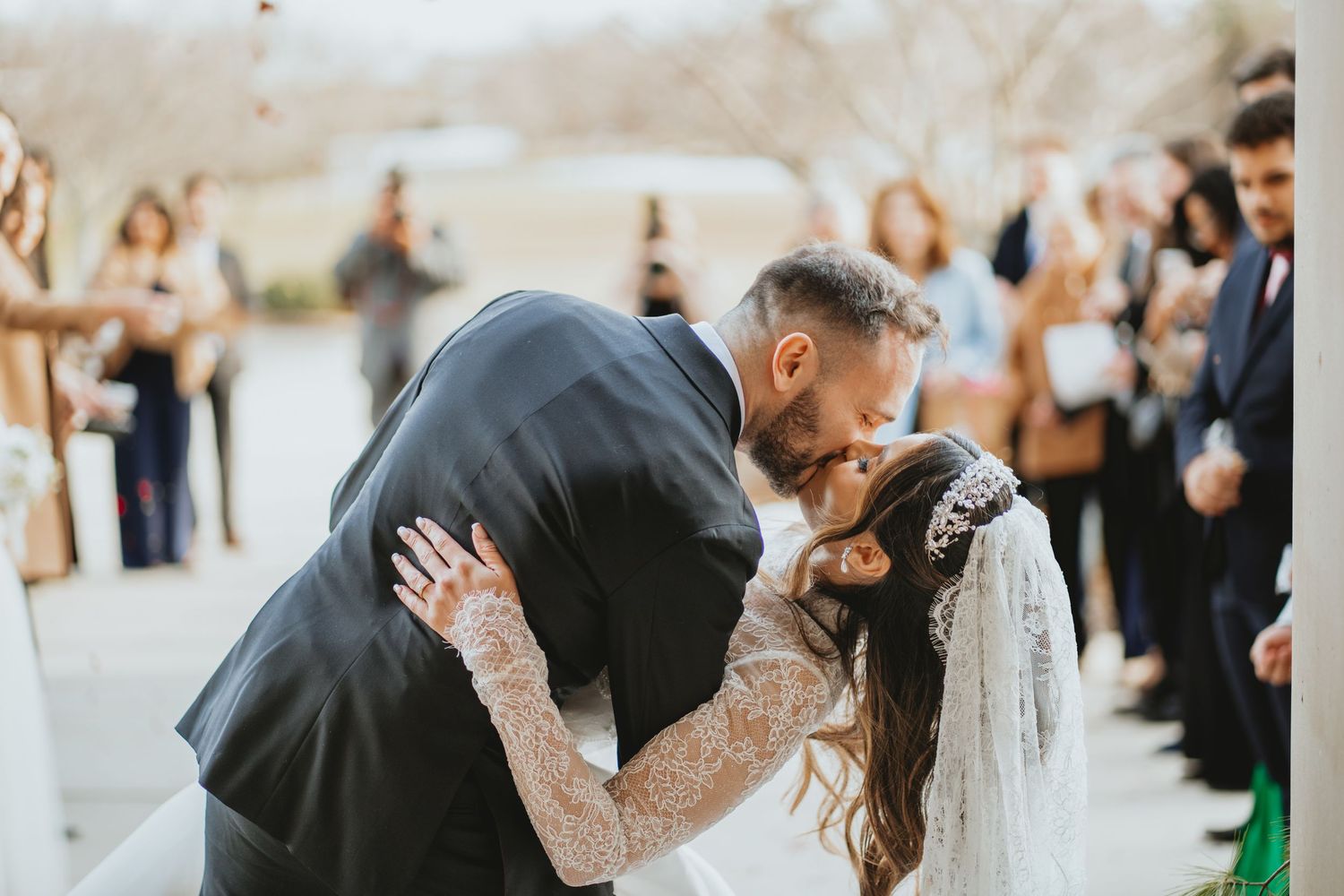 Bride and groom share romantic first kiss outdoors during wedding ceremony with guests applauding.