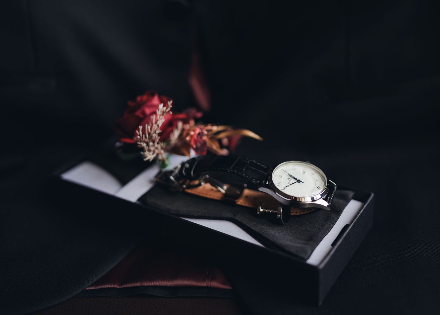 Groom's accessories flat lay with a classic white-dial watch and dried florals on dark background.