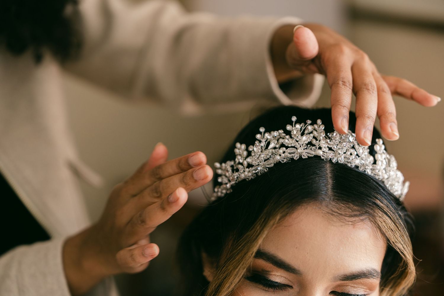 A stylist carefully places an ornate crystal and pearl bridal tiara on the bride's dark hair.