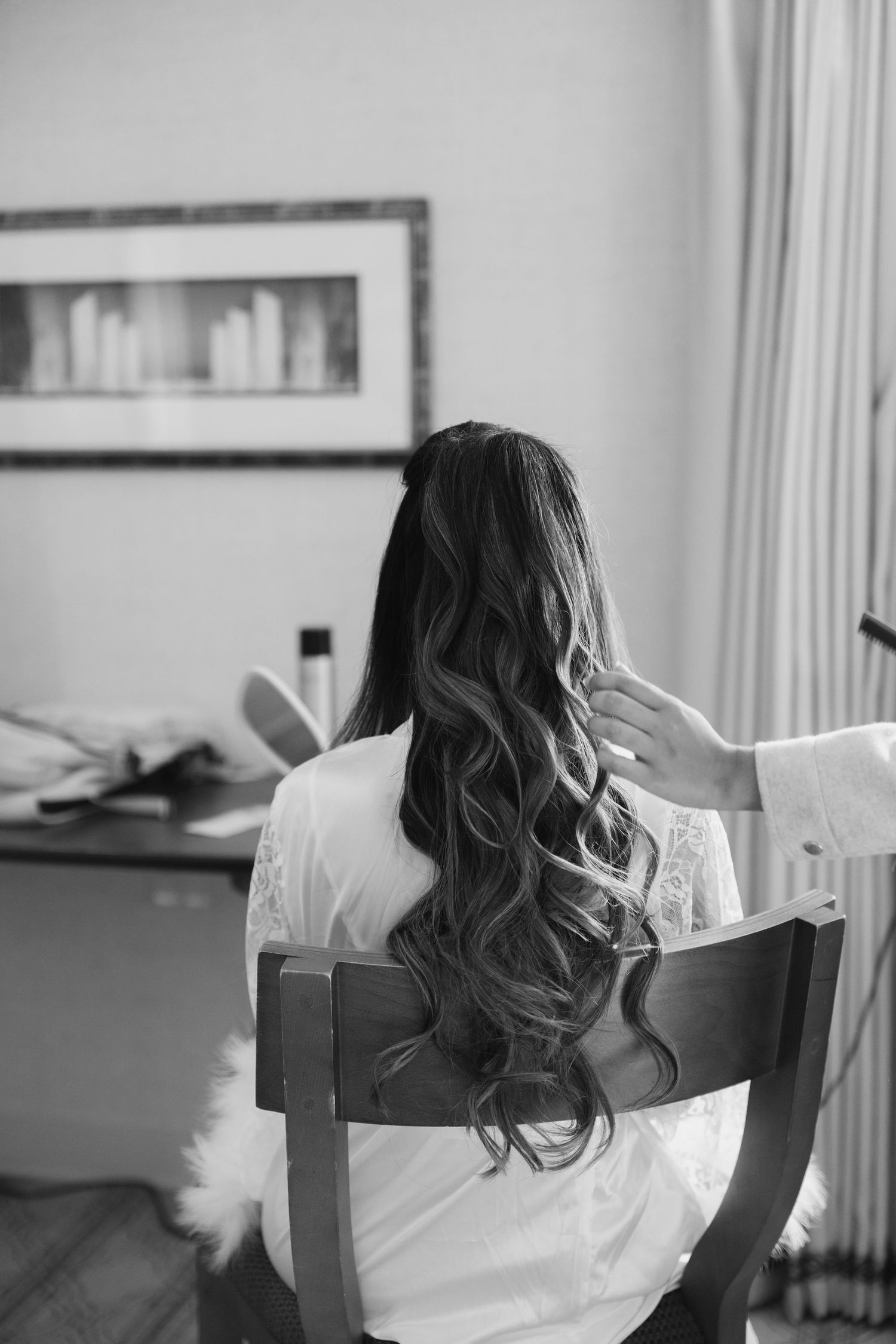 Black and white photo of a bride with long curly hair being styled while seated in a chair.