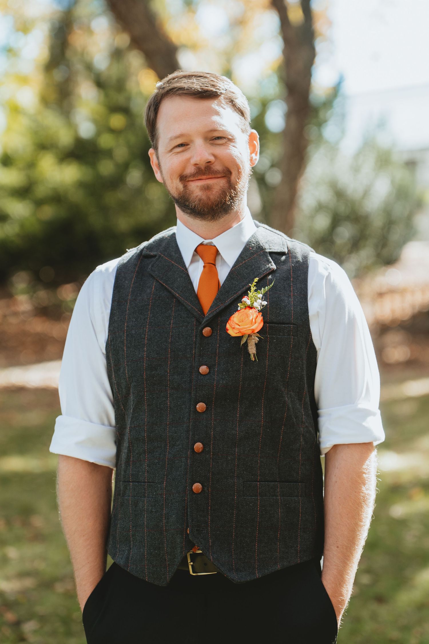 Smiling groom in tweed vest, orange tie, and boutonniere poses outdoors on his wedding day.