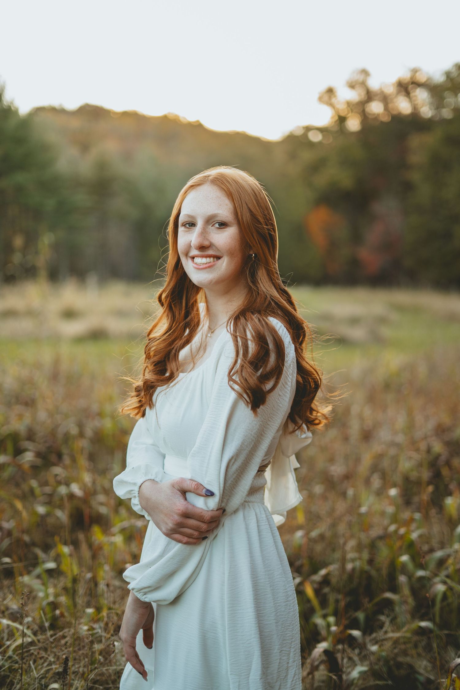 Beautiful redhead girl in white dress smiling in a golden autumn meadow at sunset.