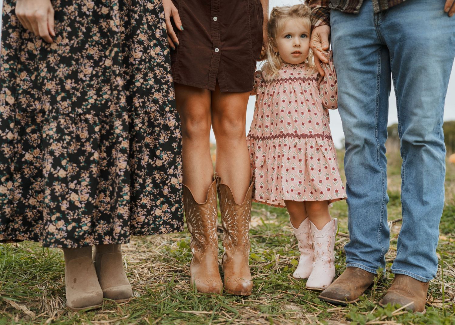 Family of three standing in field, toddler girl in floral dress with cowboy boots.