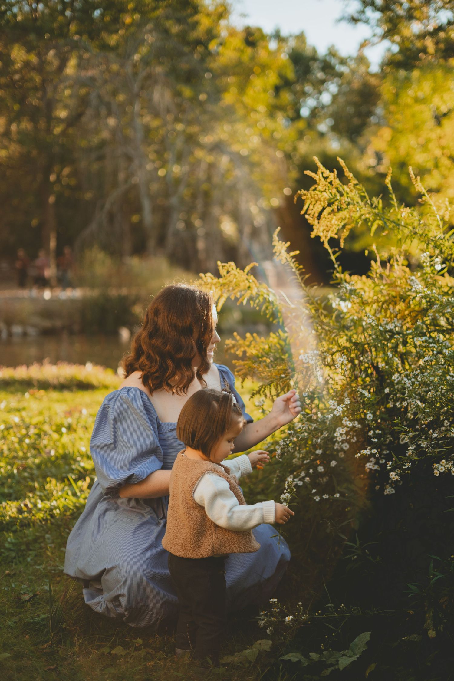 Mother and toddler daughter exploring wildflowers together in a sunlit autumn garden near a pond.