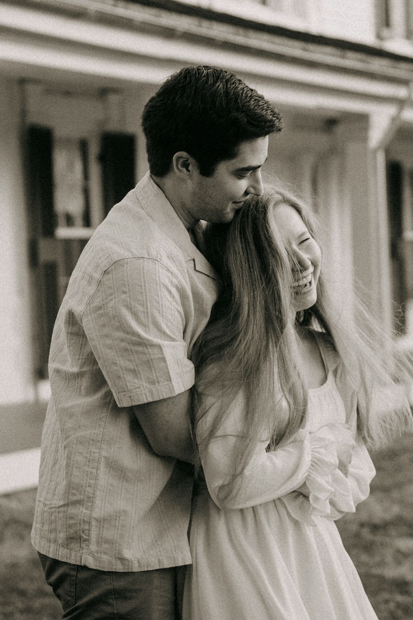 A man embraces a laughing woman from behind in a tender black and white outdoor couple portrait.