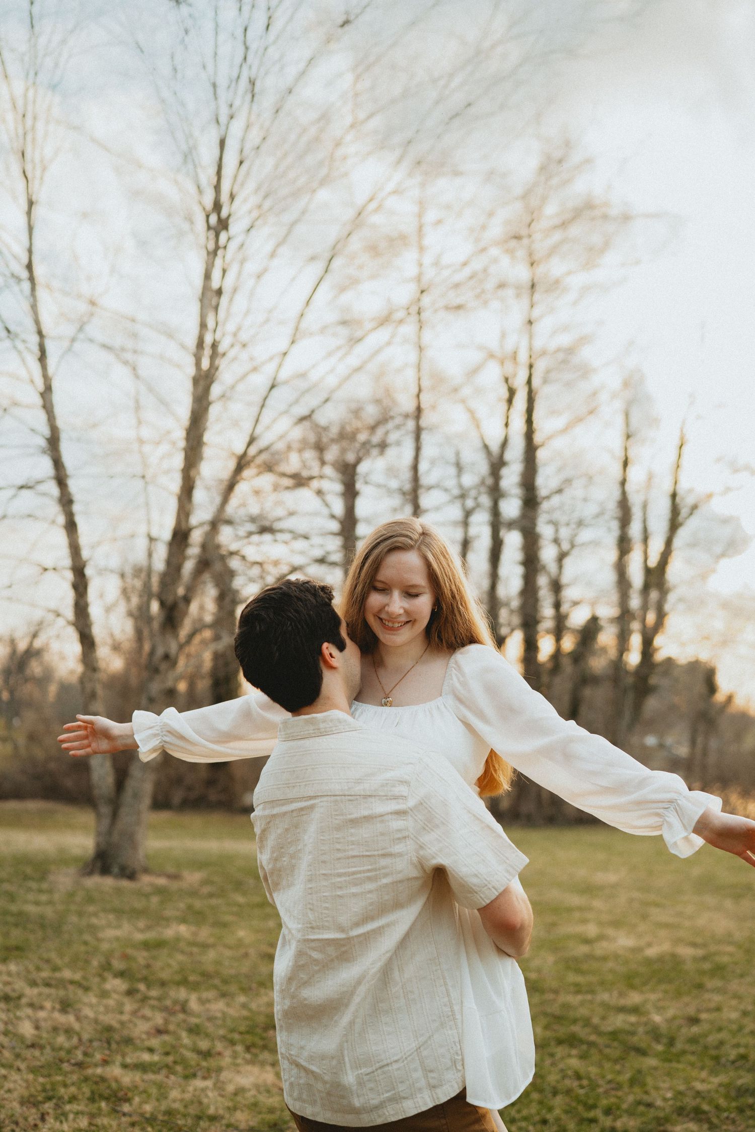 Man lifts smiling woman in white dress outdoors with bare trees and warm sunset sky behind them.