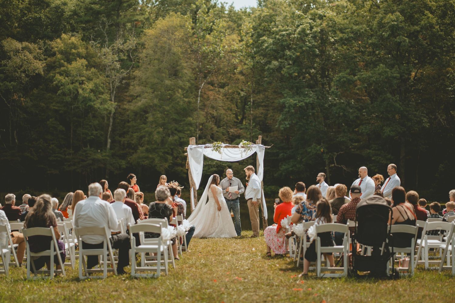 Outdoor wedding ceremony in a forest clearing with guests seated in white chairs facing a rustic floral arch.