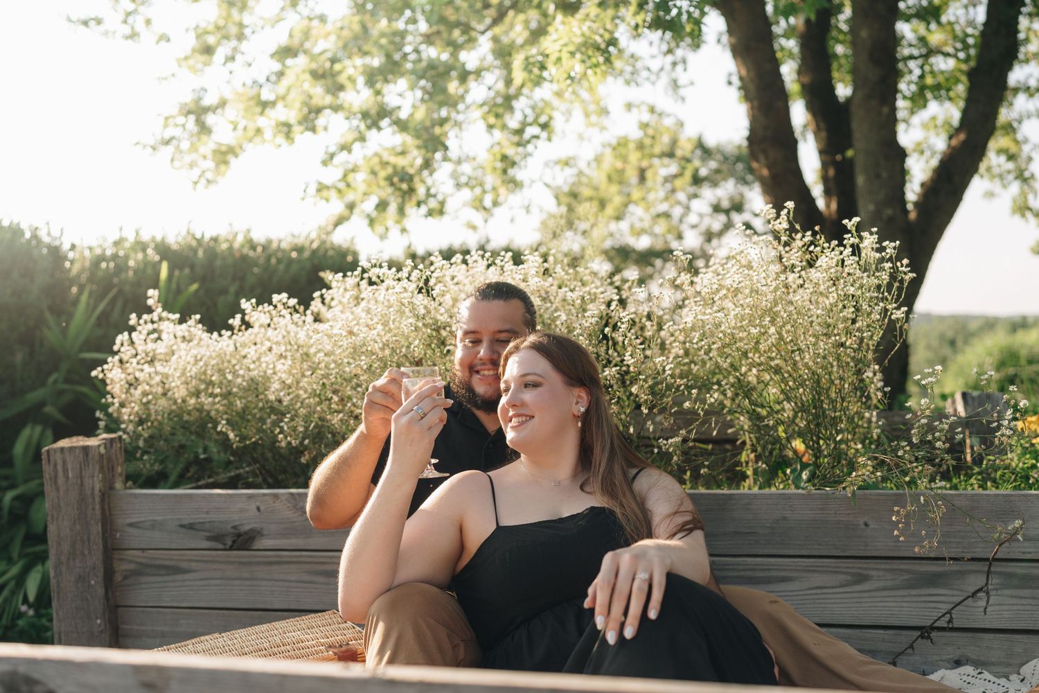 Smiling couple lounging on a wooden bench in a sunny wildflower garden setting.