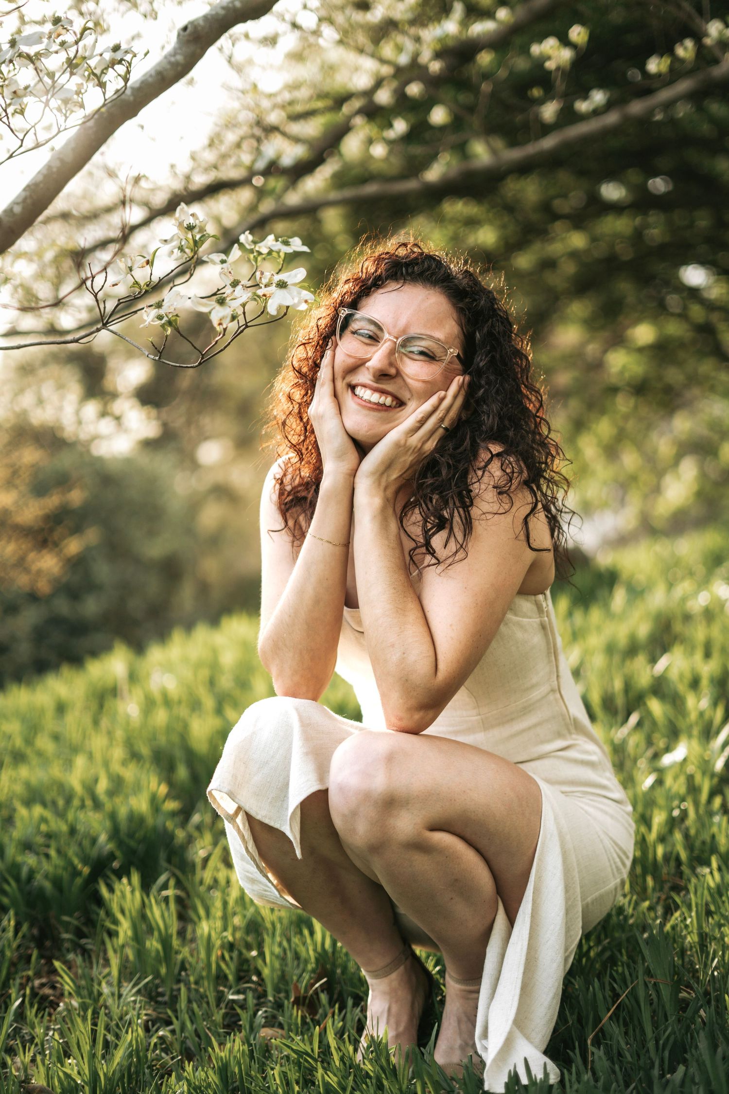 Joyful curly redhead woman in white dress crouching on grass, smiling with hands on cheeks.