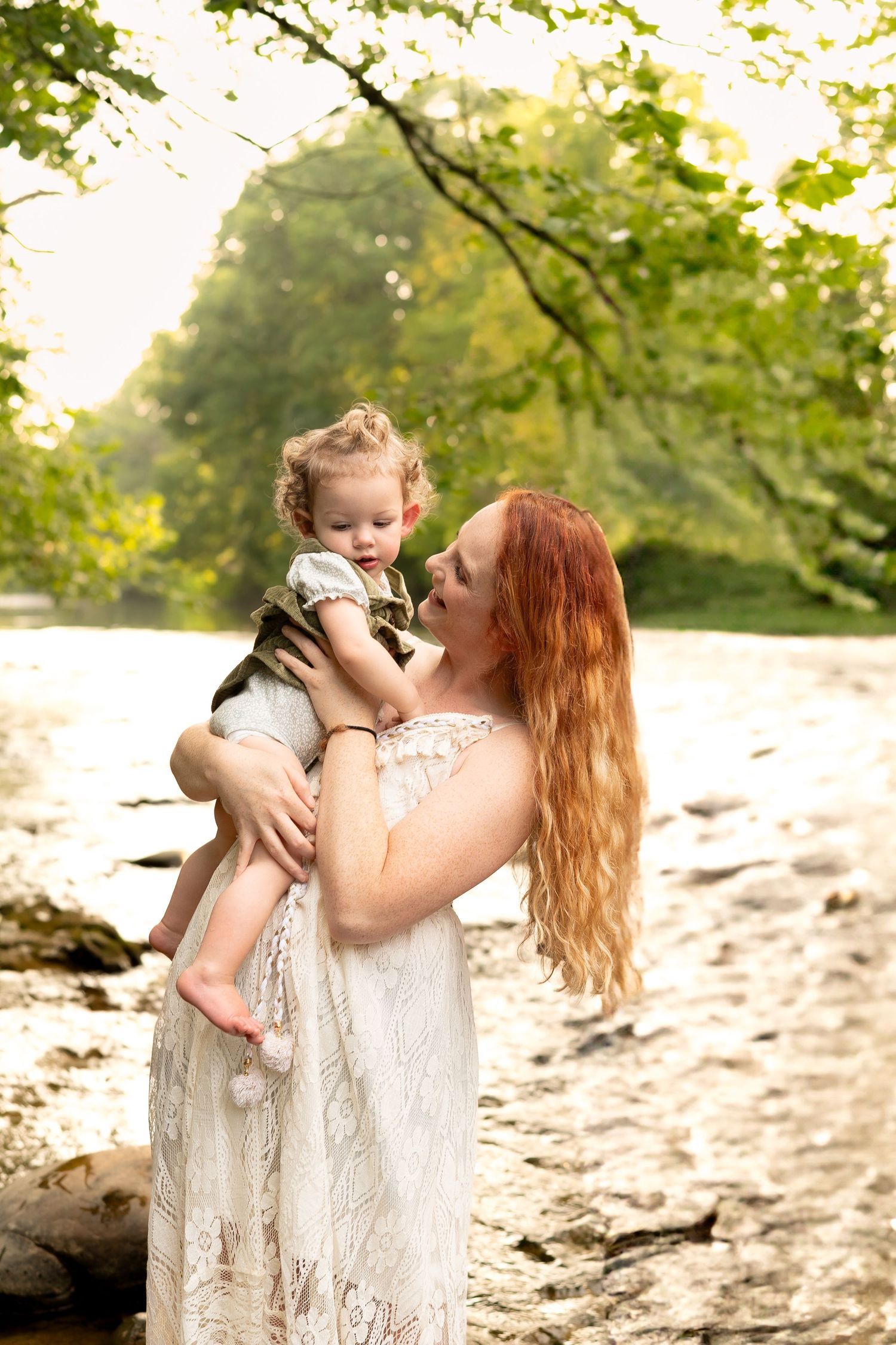 Red-haired mother in white lace dress tenderly holding her toddler by a sunlit riverside.