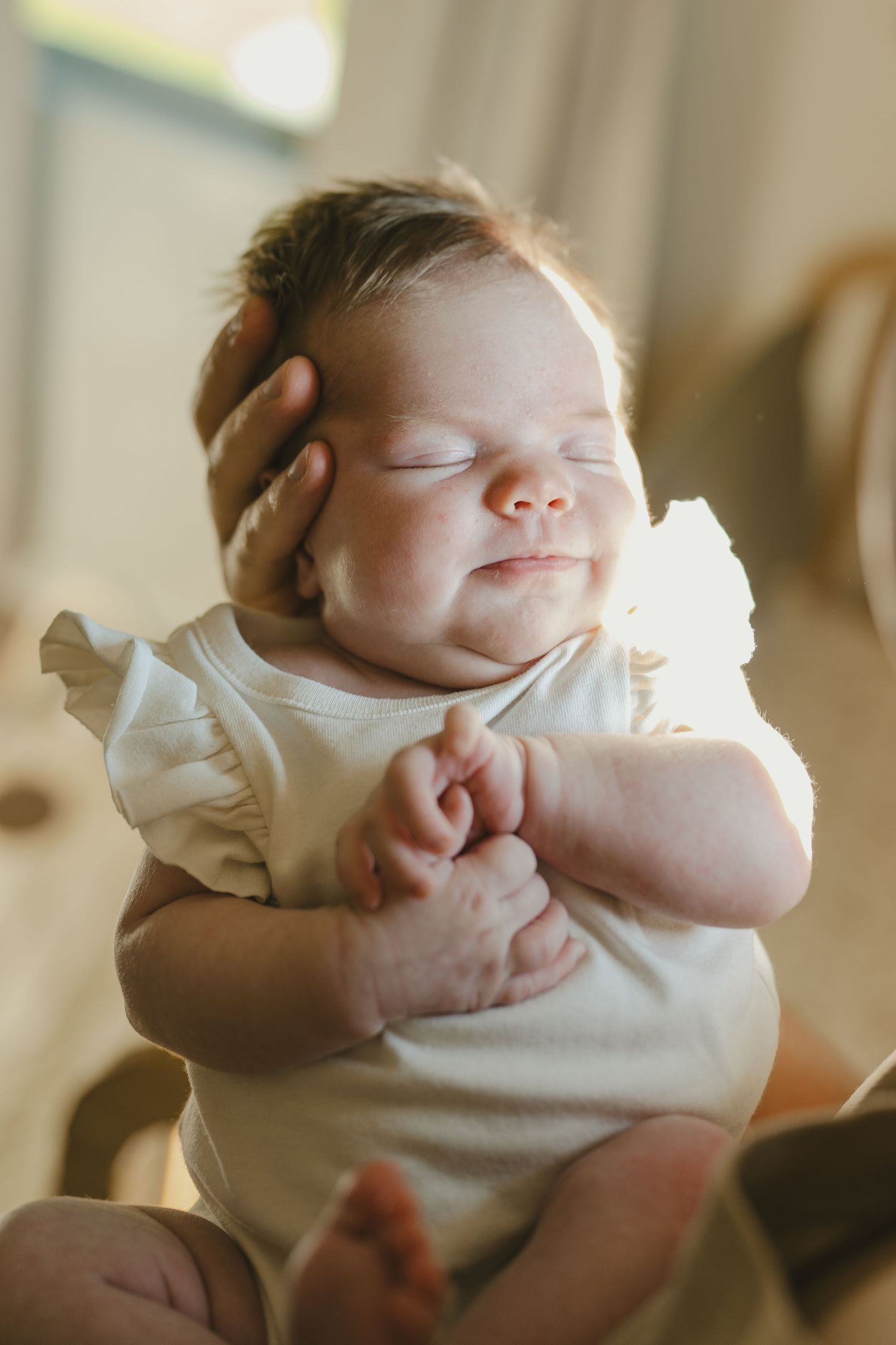 Peaceful sleeping newborn baby girl in white ruffle onesie held gently in parent hands.