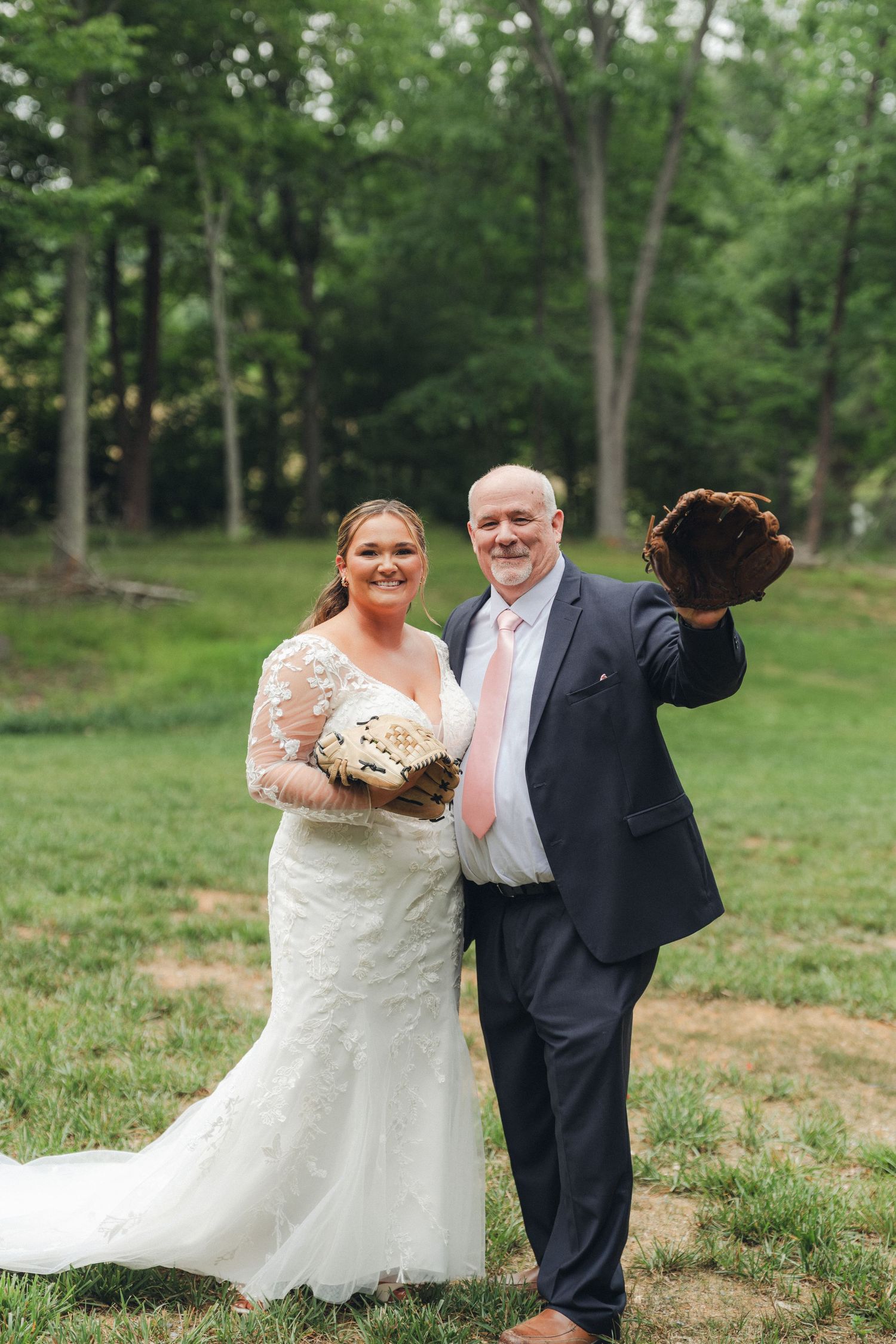 Bride and groom smiling outdoors, groom holding baseball glove, bride with bouquet, lush green trees background.